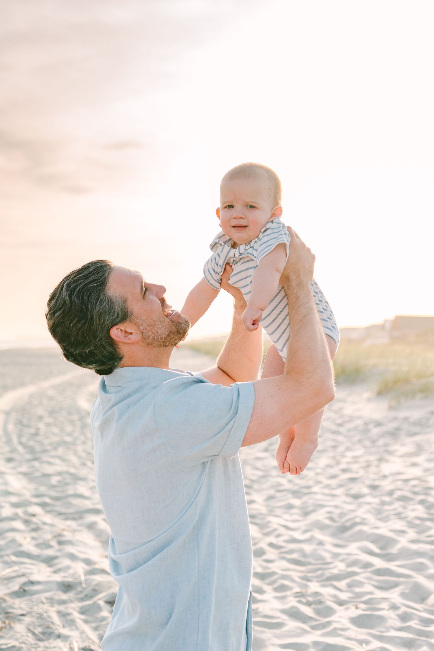 Parent lifting smiling baby against sunset beach backdrop.