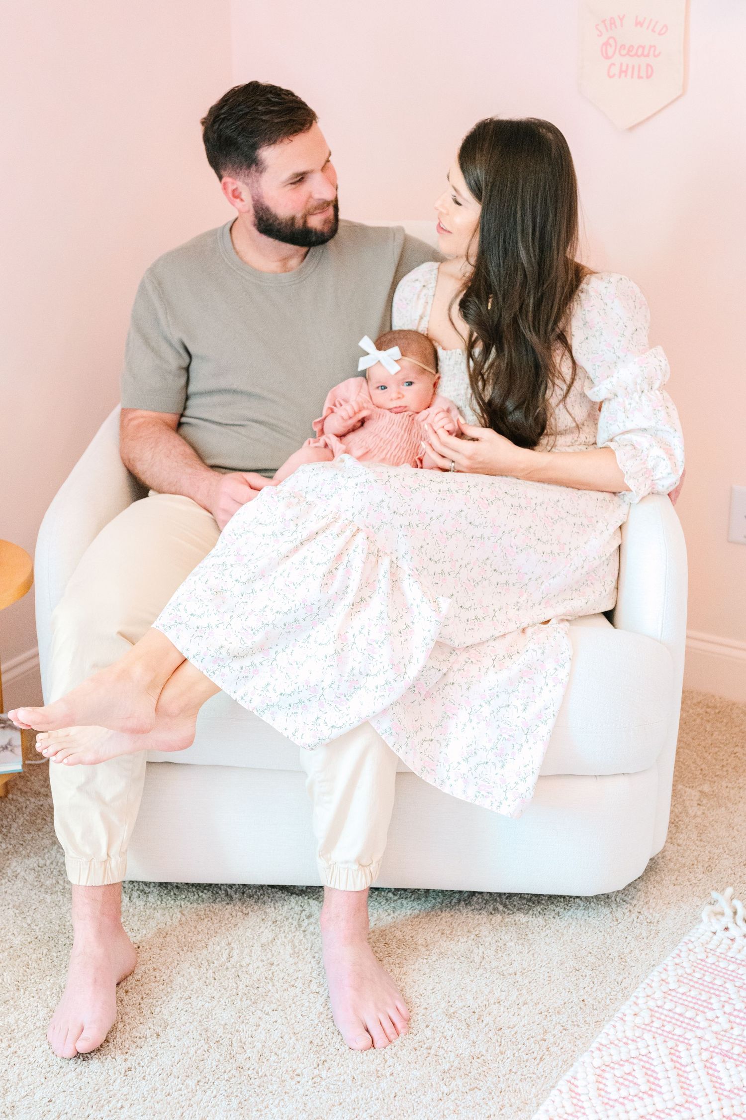 A couple sits together on a white chair while holding a baby in a pink outfit against a pink wall.