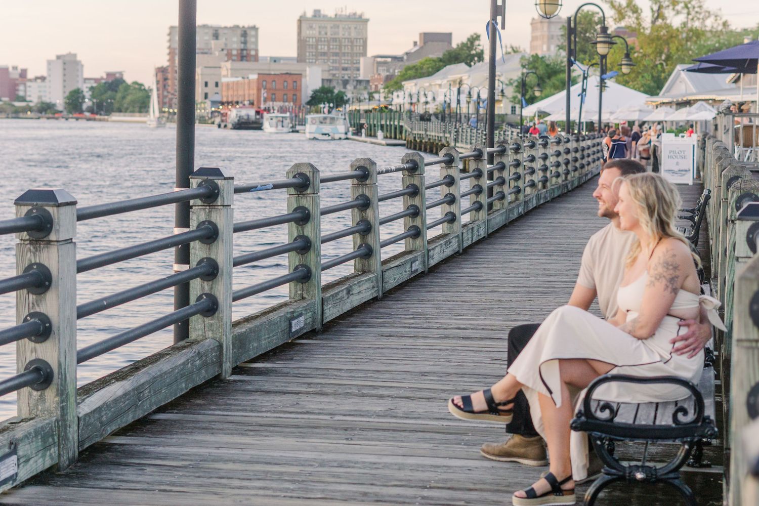 Woman in a dress relaxes on a bench along a waterfront boardwalk on a sunny day.