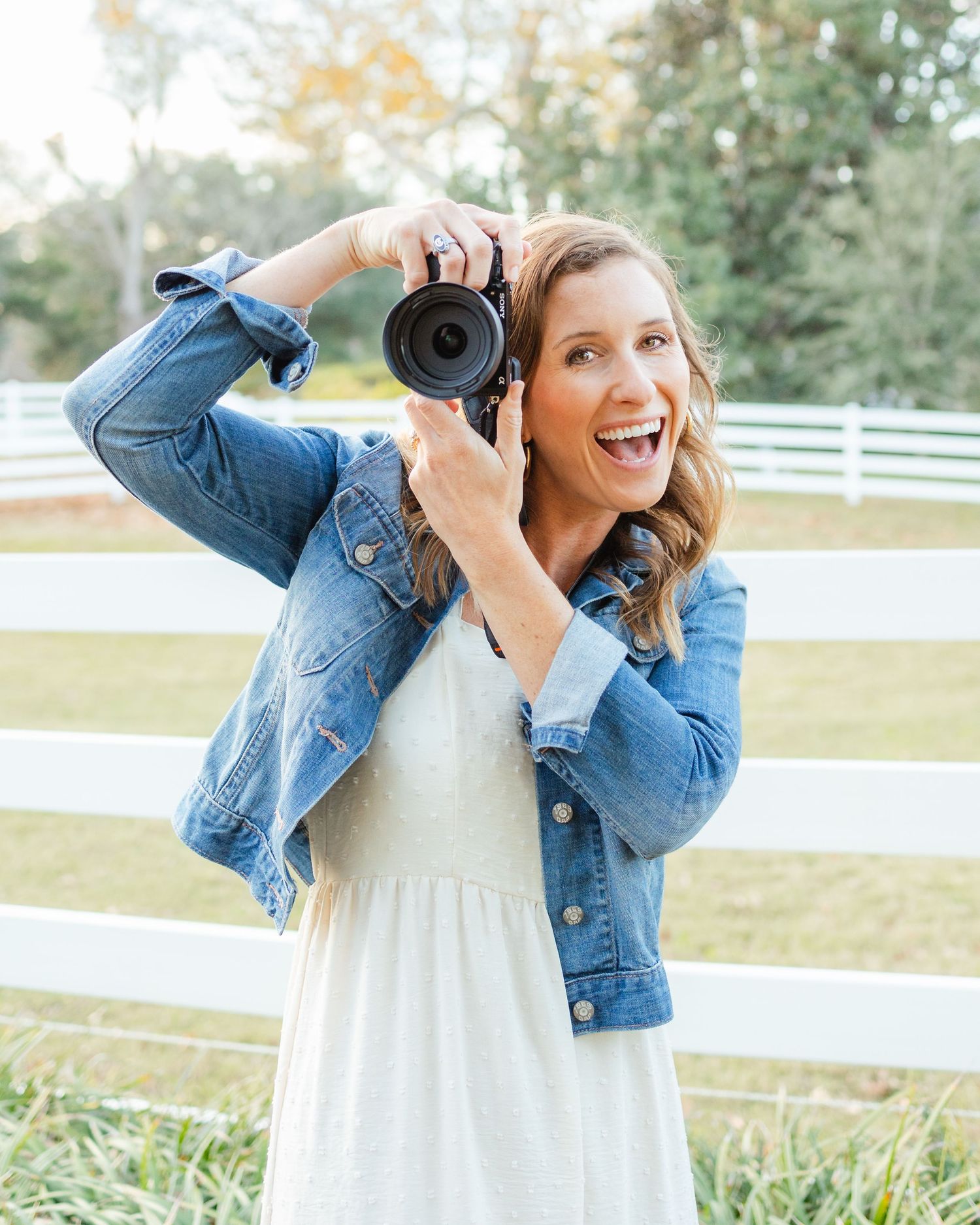 Photographer in denim jacket and white dress smiles while holding camera against white fence backdrop.