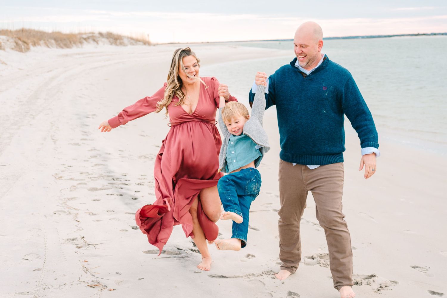 Family walking and playing together on a sandy beach with water and sand dunes in background.