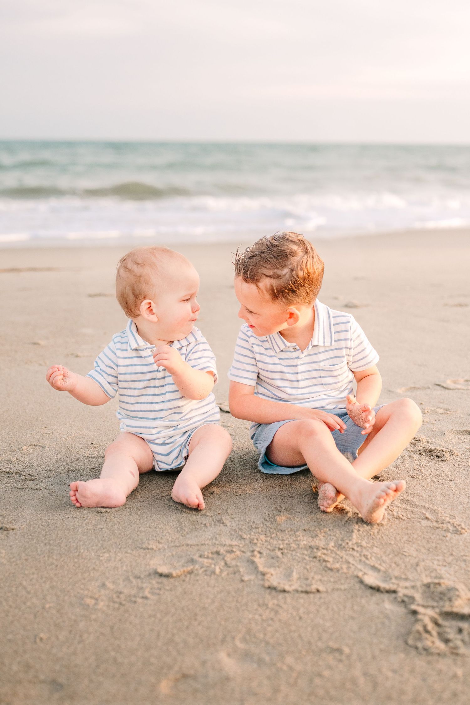 Two young children in striped outfits sitting together on beach sand.