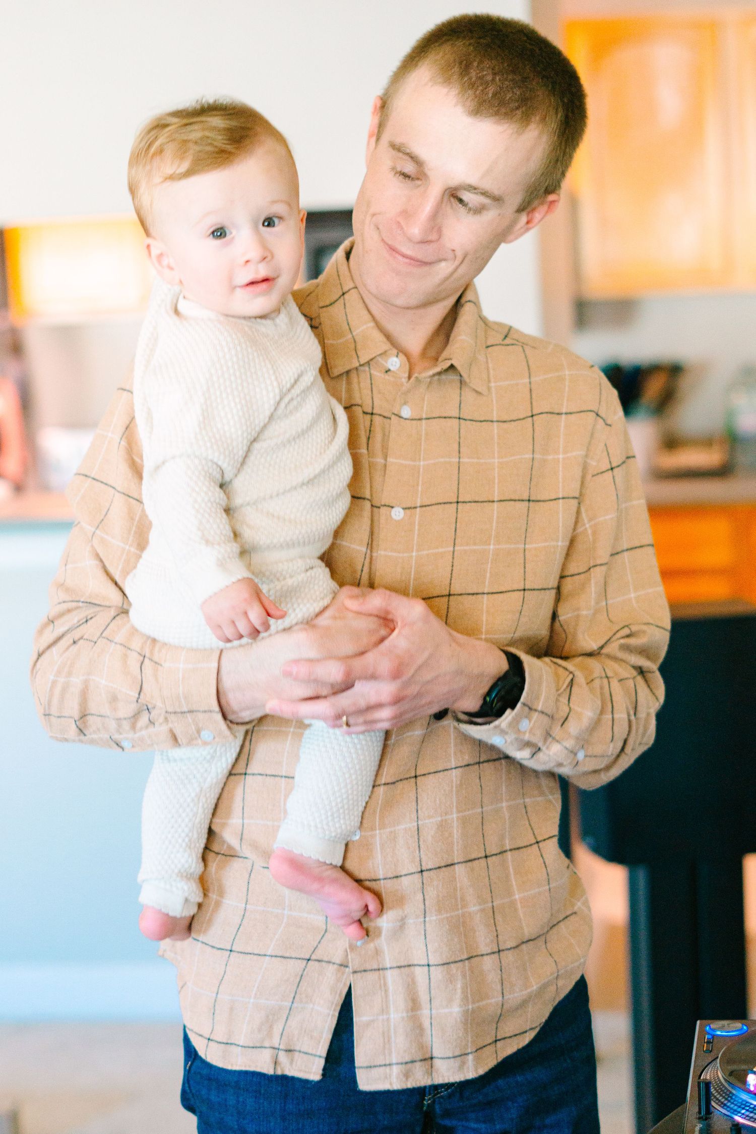 Parent holding infant in plaid shirt and jeans in bright kitchen setting.