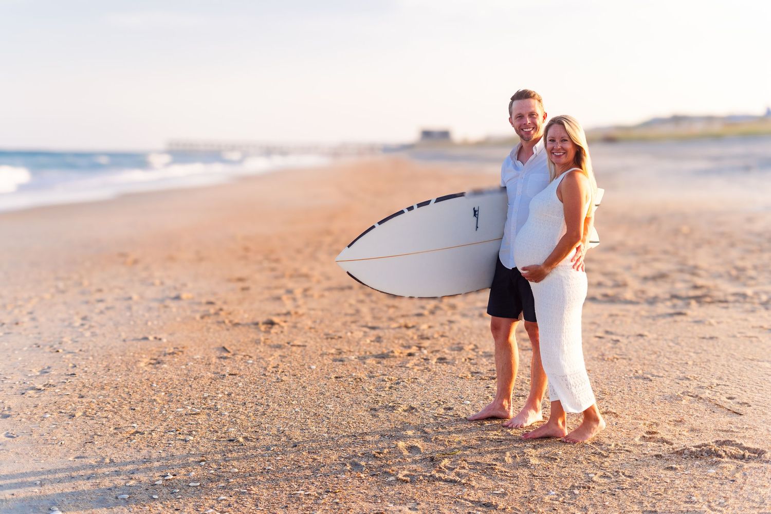 Expectant parents stand together on the beach holding a surfboard during golden hour.