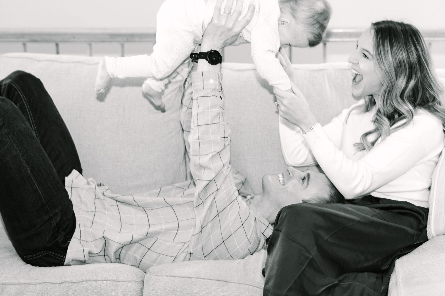 Parents playing with baby on white couch in black and white photo.
