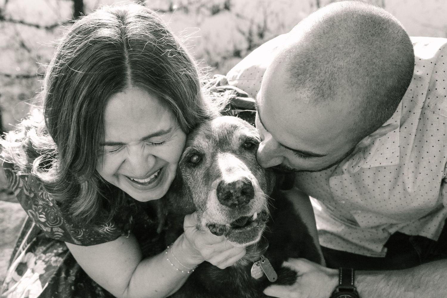 A black and white photo of a couple cuddling with their smiling dog.