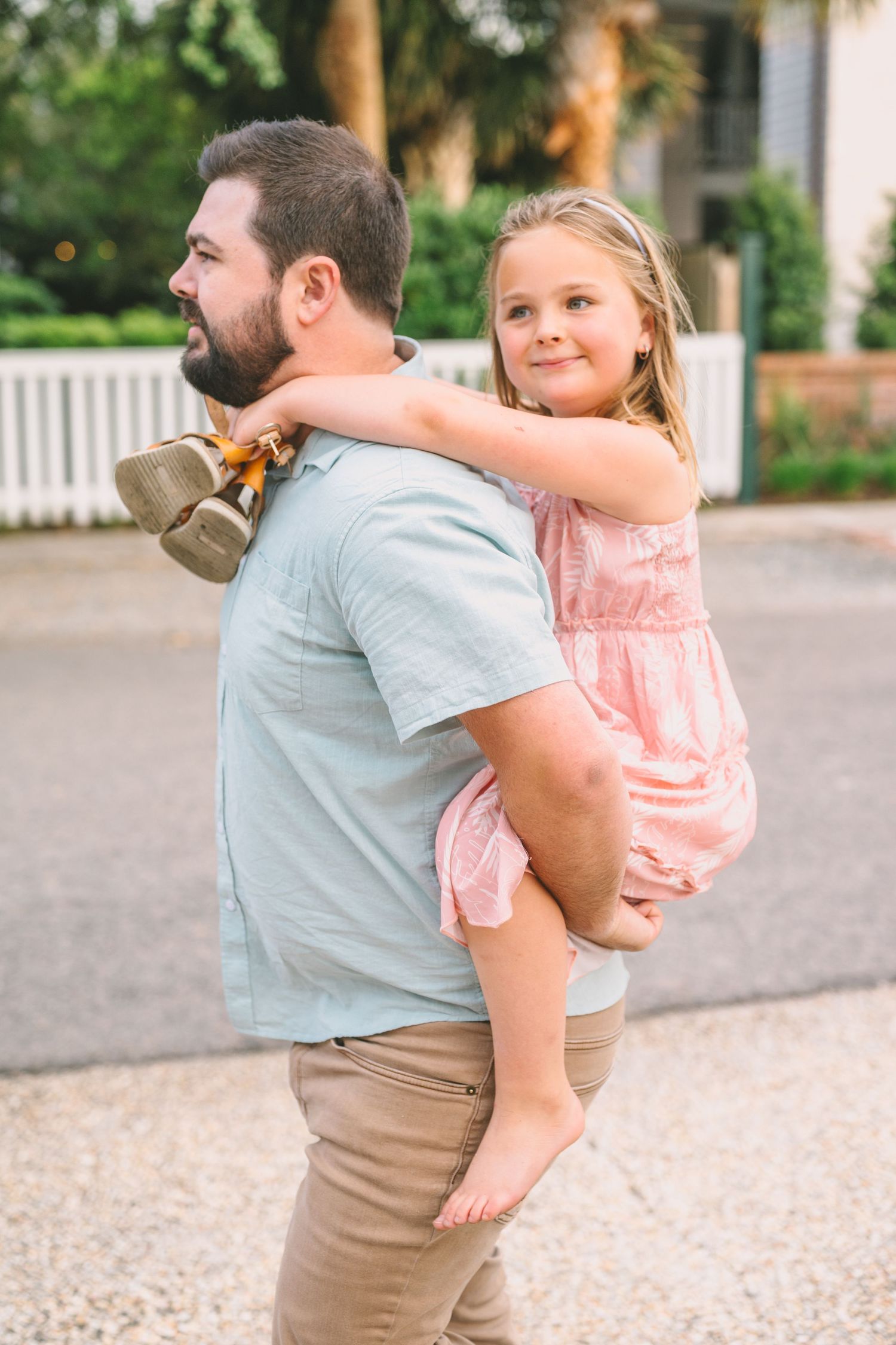 A young child in a pink dress rides piggyback on an adult's shoulders as they smile together outdoors.