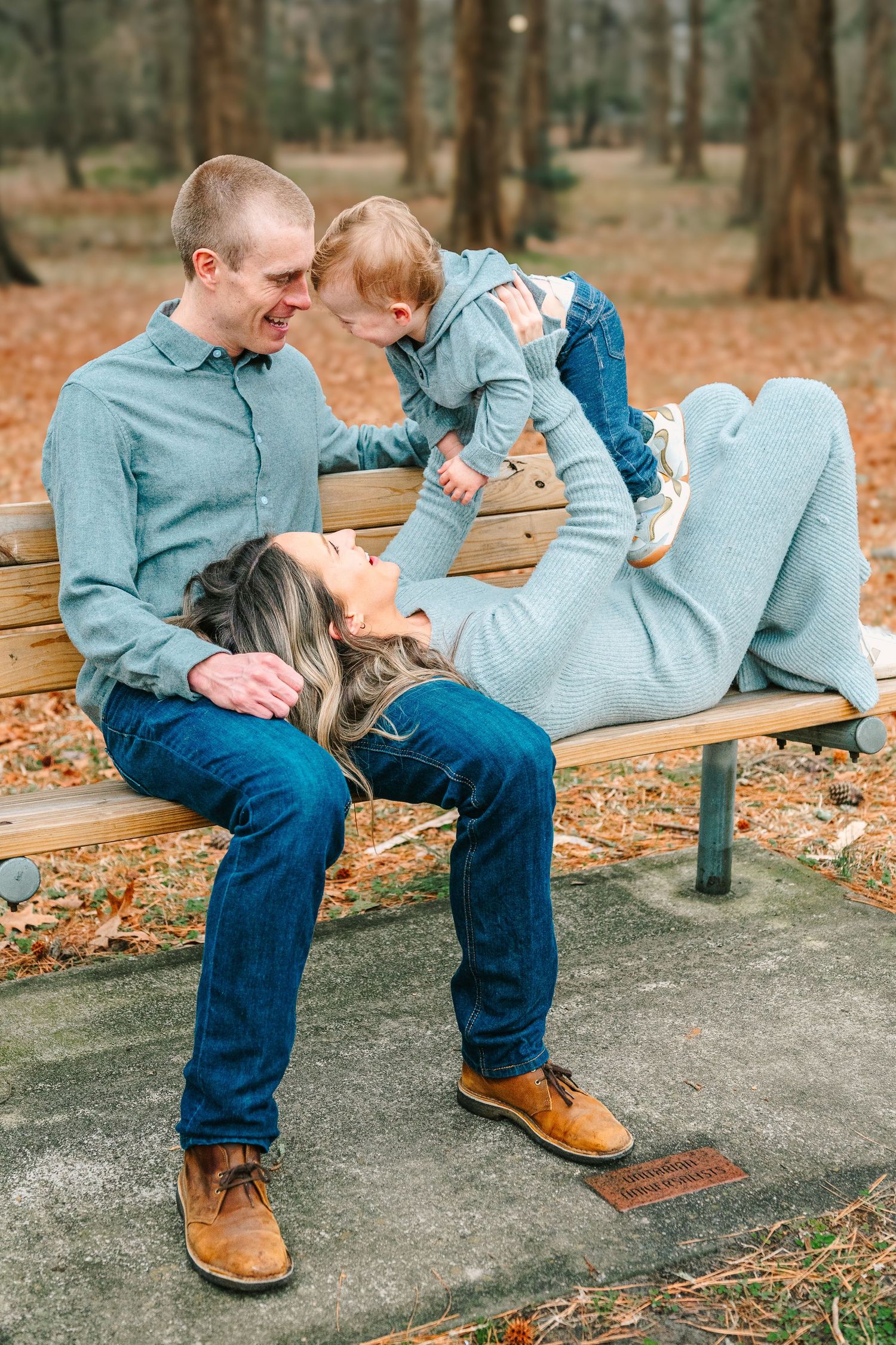 Family enjoys playful moment together on park bench surrounded by autumn leaves and tall pine trees.