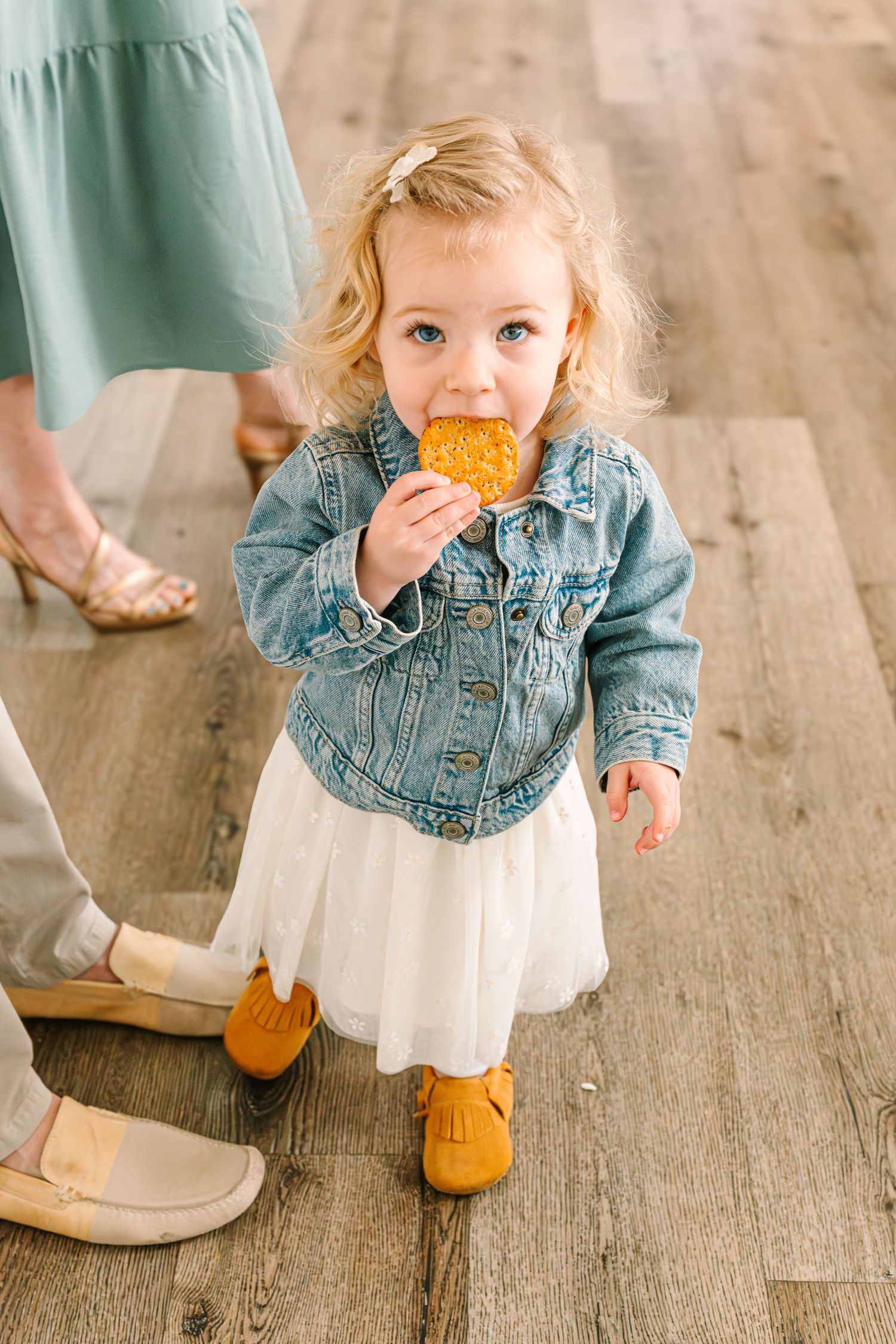 A small child in a denim jacket and white dress enjoys eating a cookie while wearing mustard yellow moccasins.