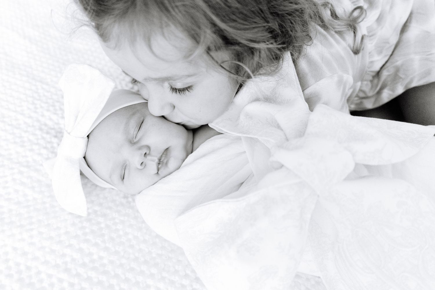 A tender black and white photo of a sleeping infant being cuddled by a toddler on white bedding.