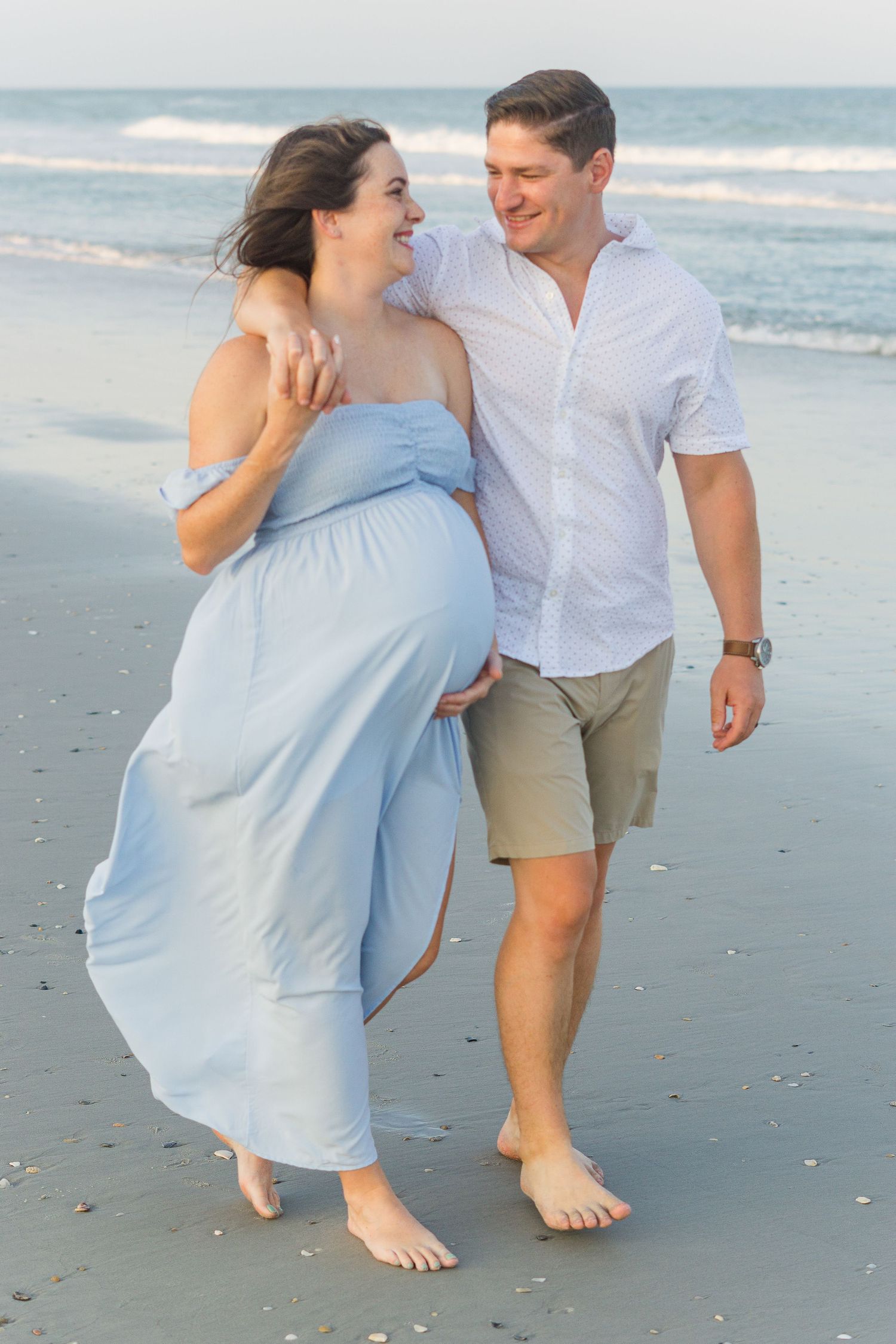 A couple walking barefoot on the beach during a maternity photo session at sunset.