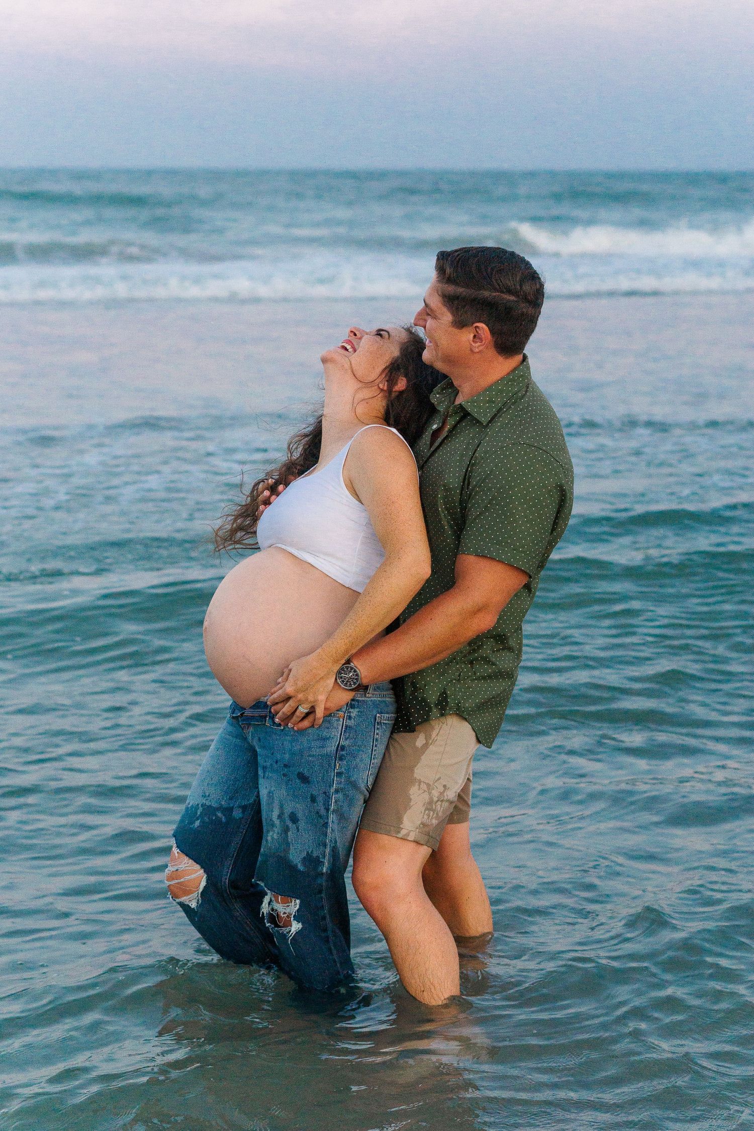 A joyful maternity moment captured as a couple stands in the ocean waves.