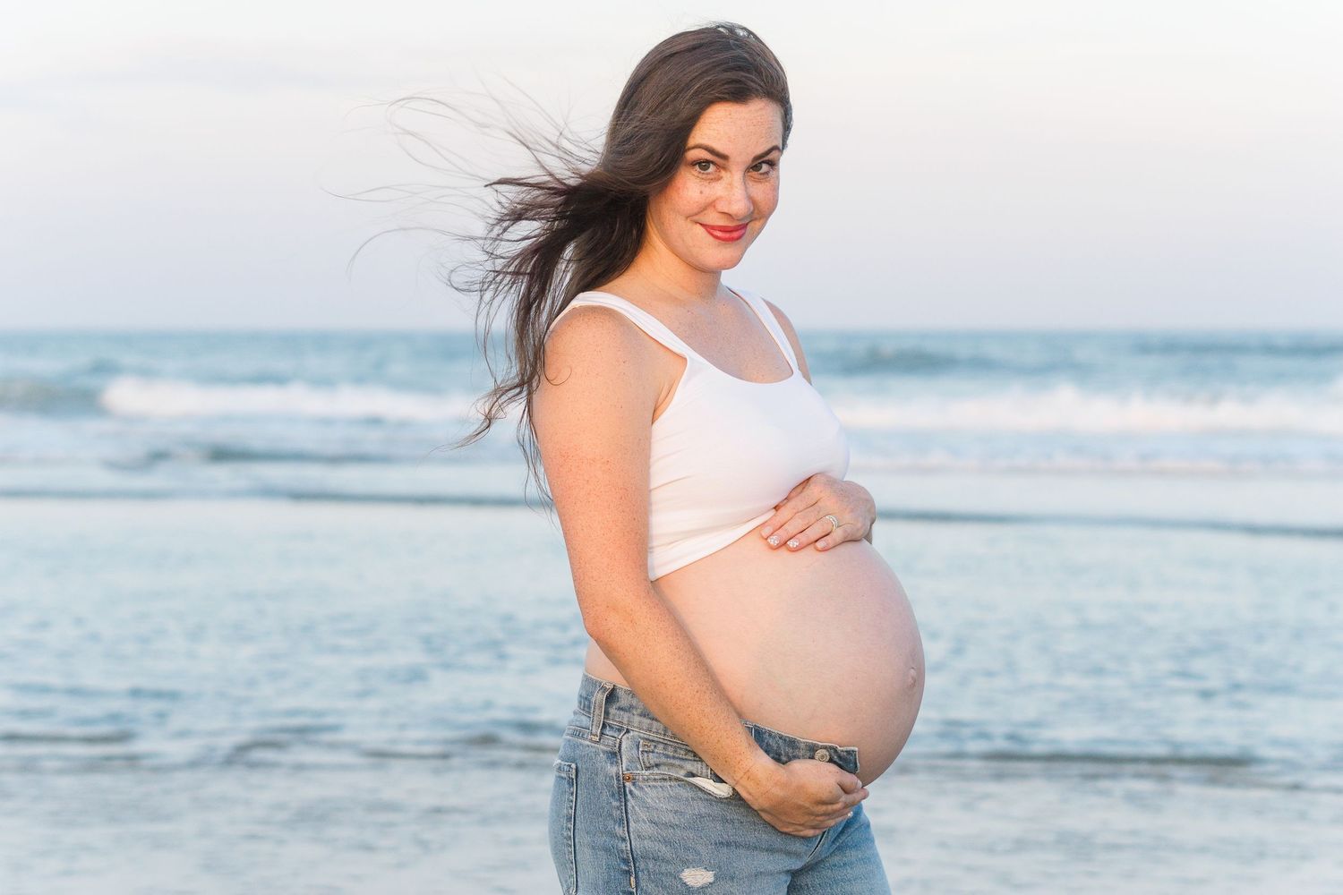 Expectant mother in casual beachwear poses with ocean waves behind her.