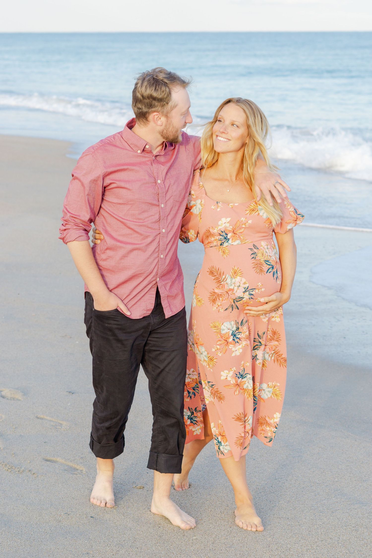A romantic couple walks barefoot along a beach shoreline at sunset.