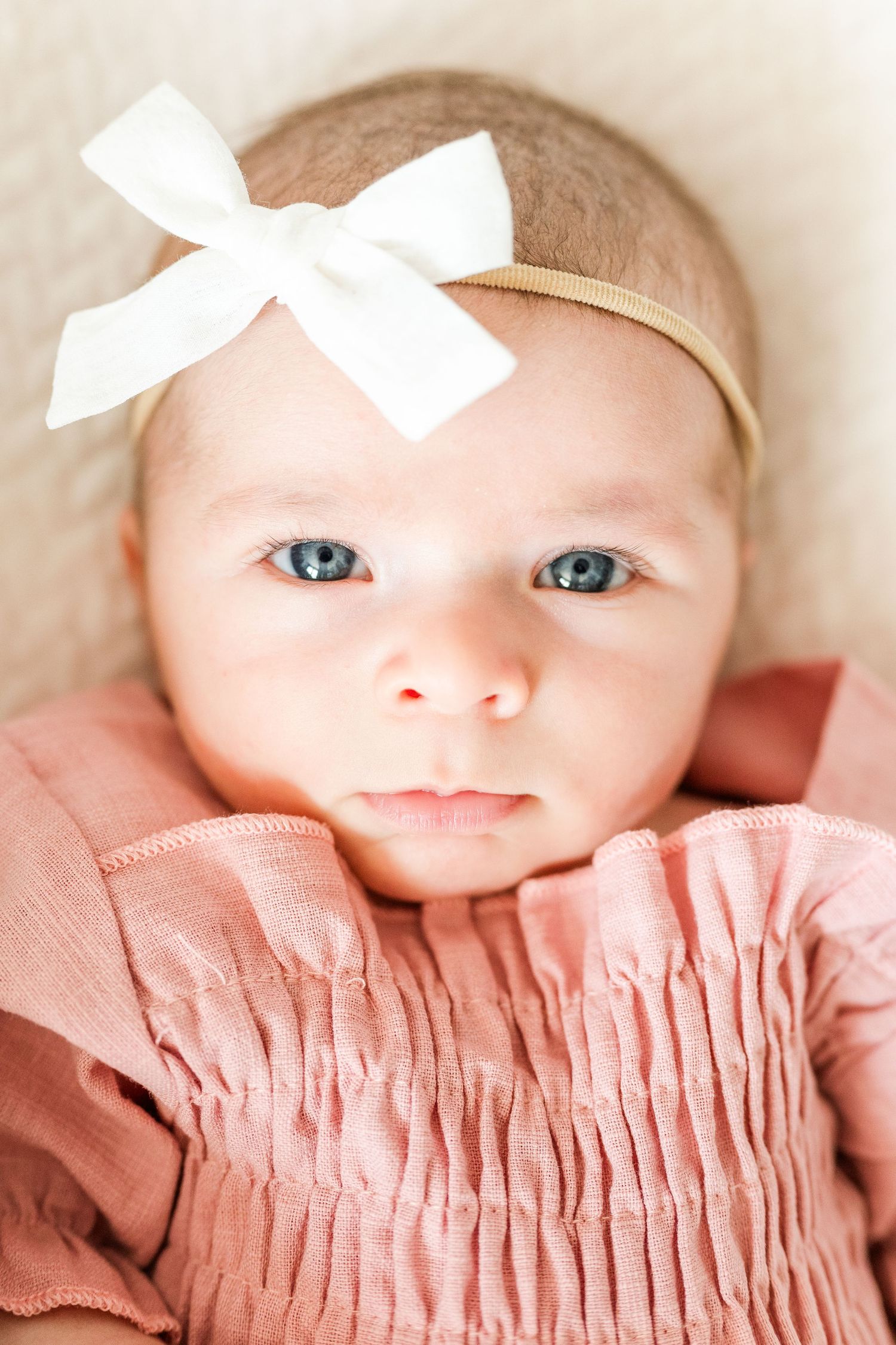 Close-up of a baby wearing a pink smocked dress and white bow headband looking directly at the camera.