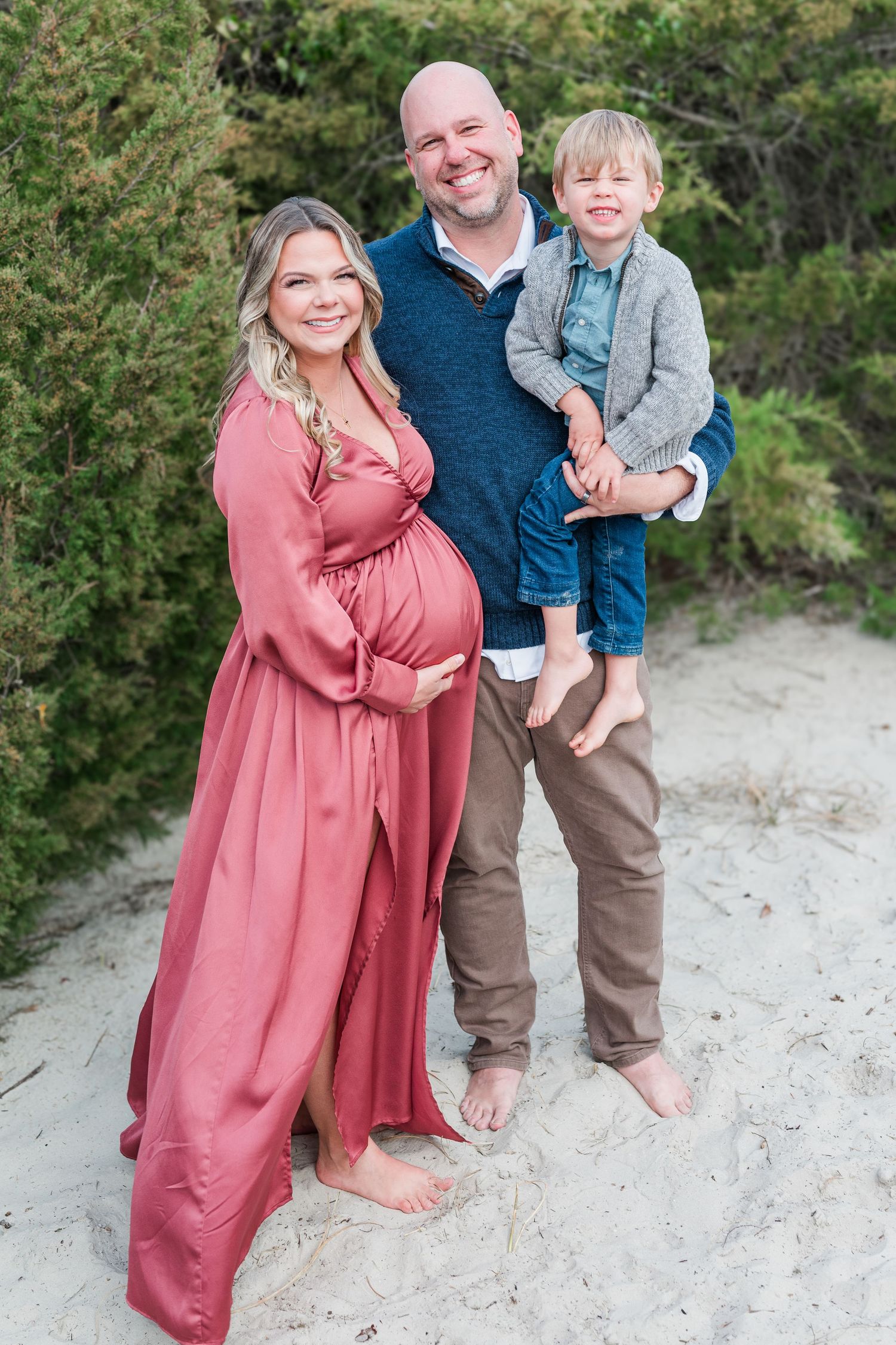 A family stands barefoot on a sandy path with trees in the background, featuring an expectant mother in a pink dress.