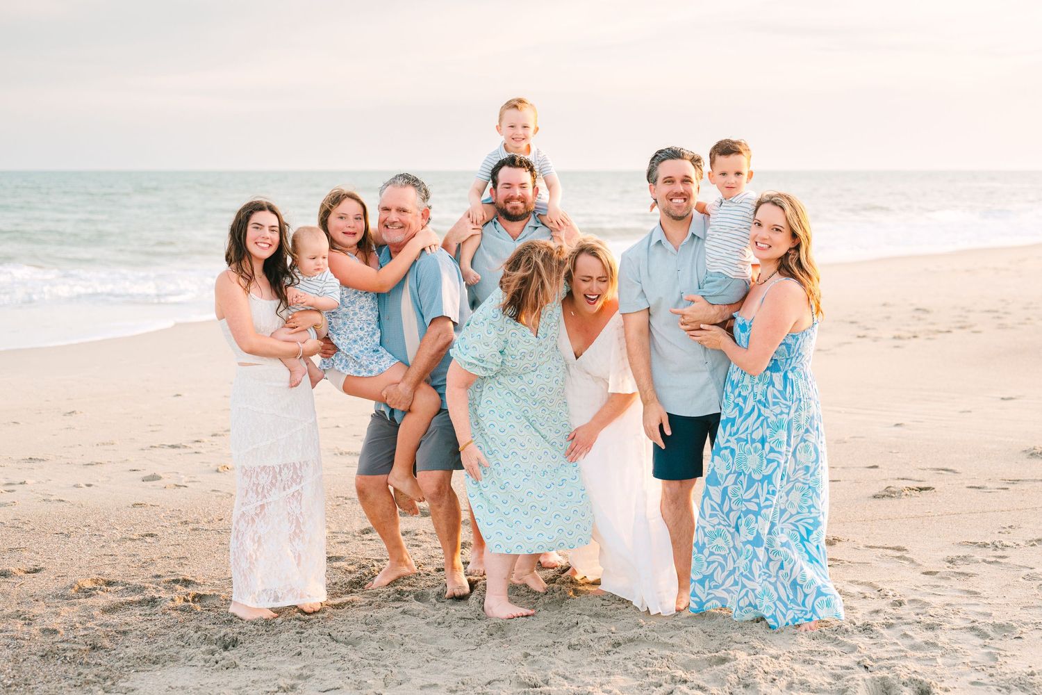 Large family group in coordinated coastal attire posing together on beach at sunset.