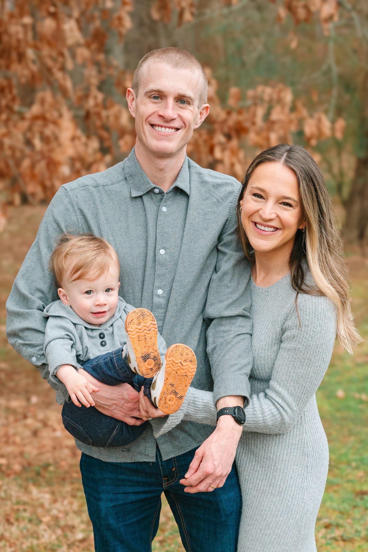 Happy family in matching grey outfits poses together outdoors during autumn with small toddler showing orange shoes.