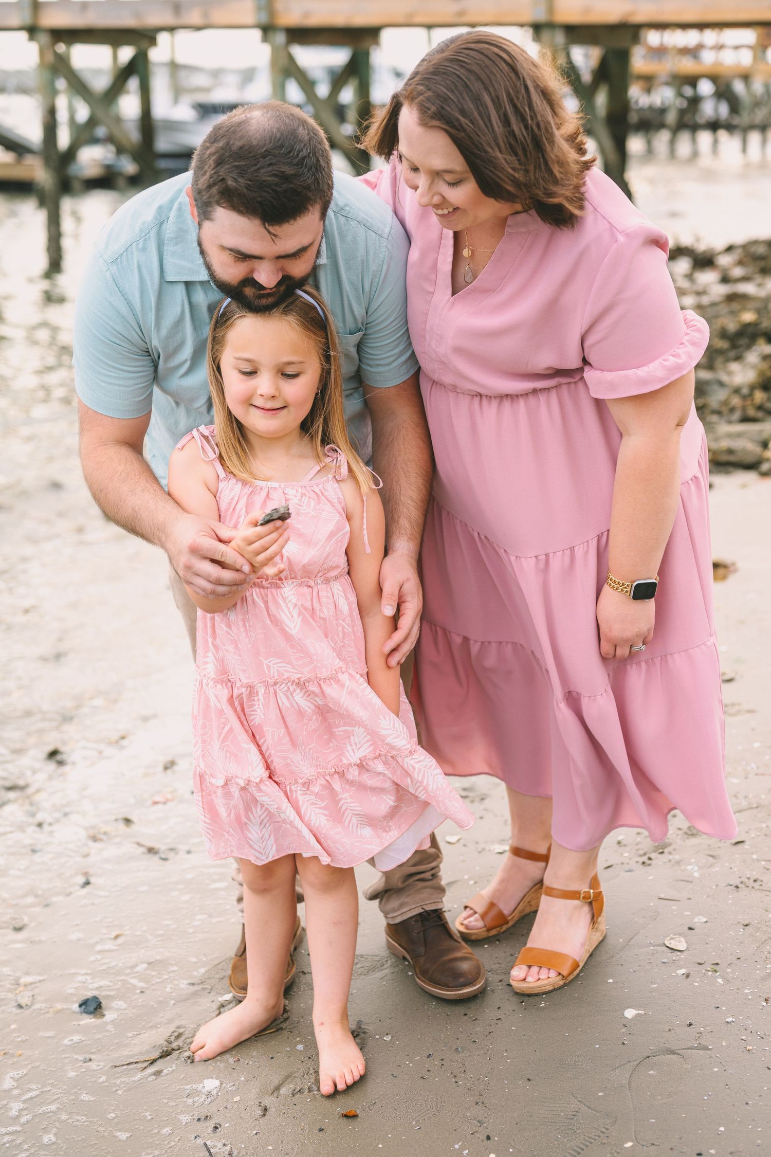 Family wearing pink and blue clothing shares a tender moment together on a sandy beach near a pier.