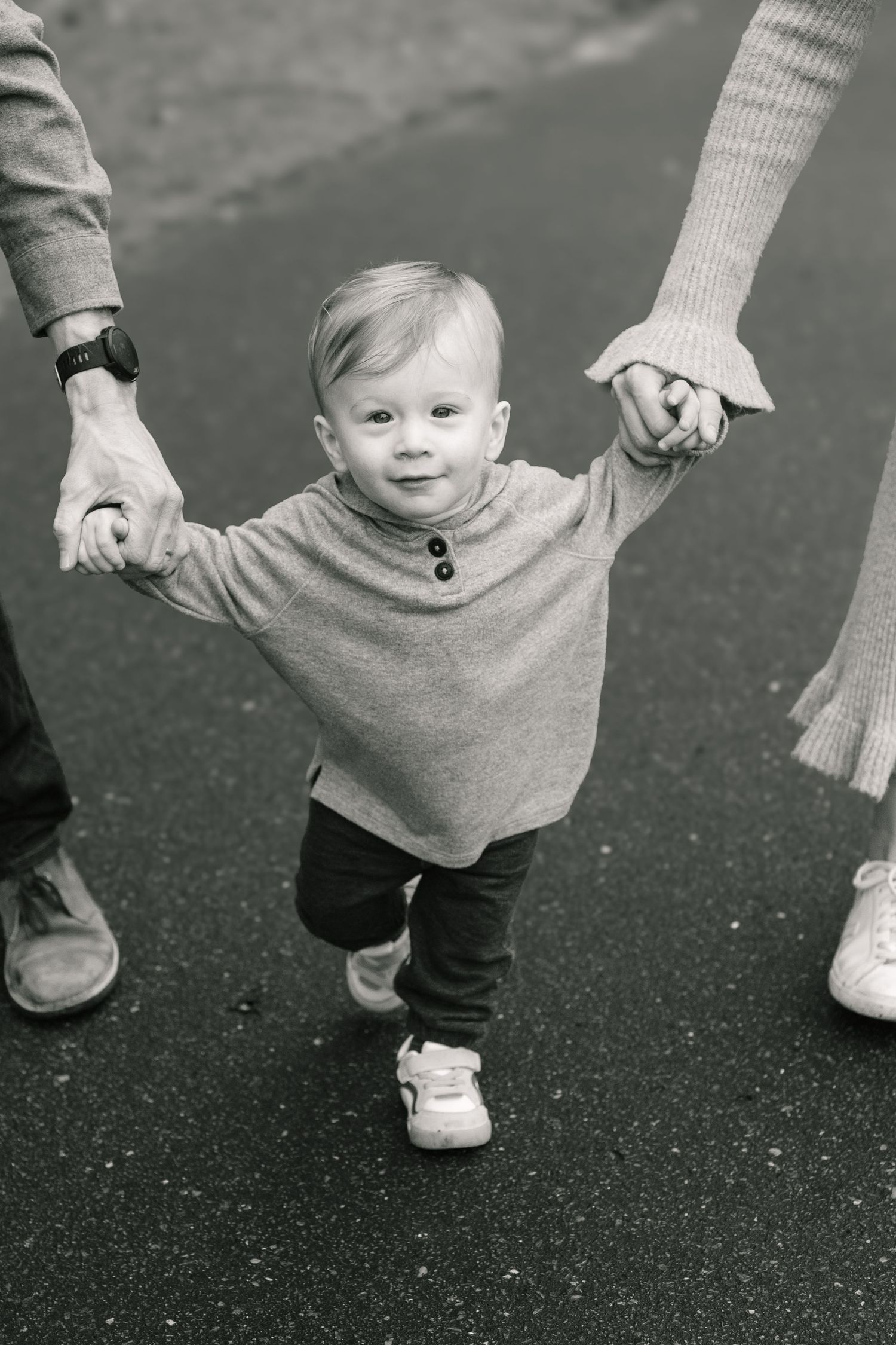 Black and white photo of toddler in sweater holding hands with adults while learning to walk on pavement.