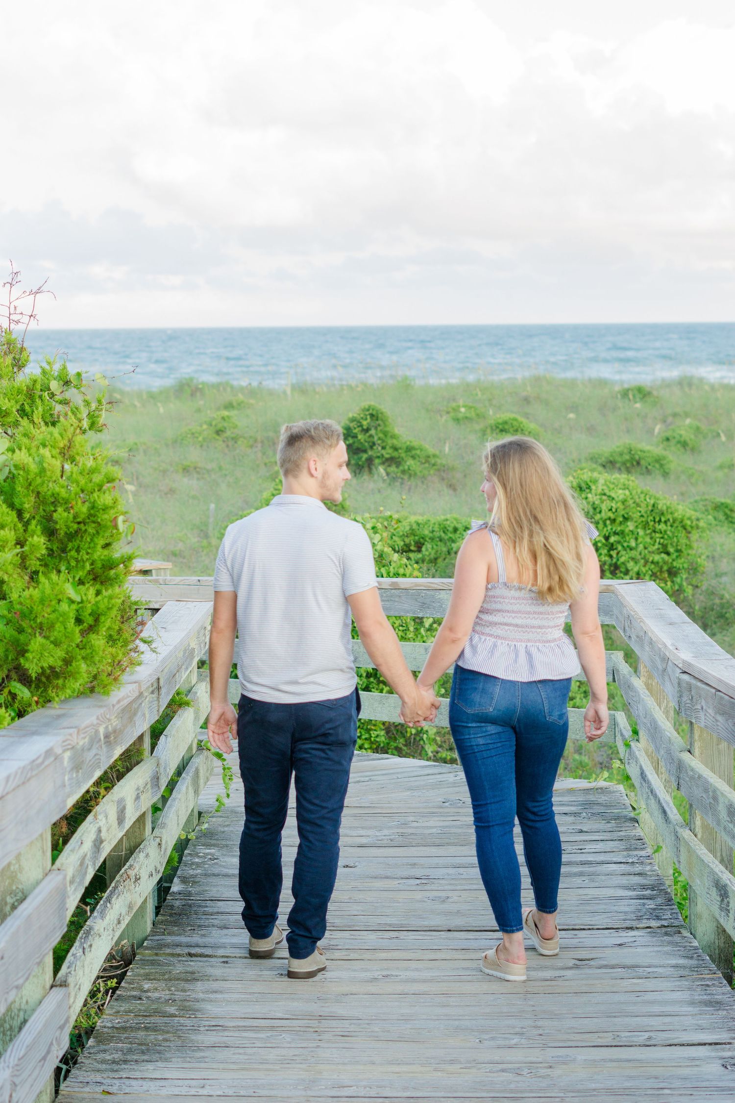 A couple holds hands while walking along a wooden boardwalk by the beach.
