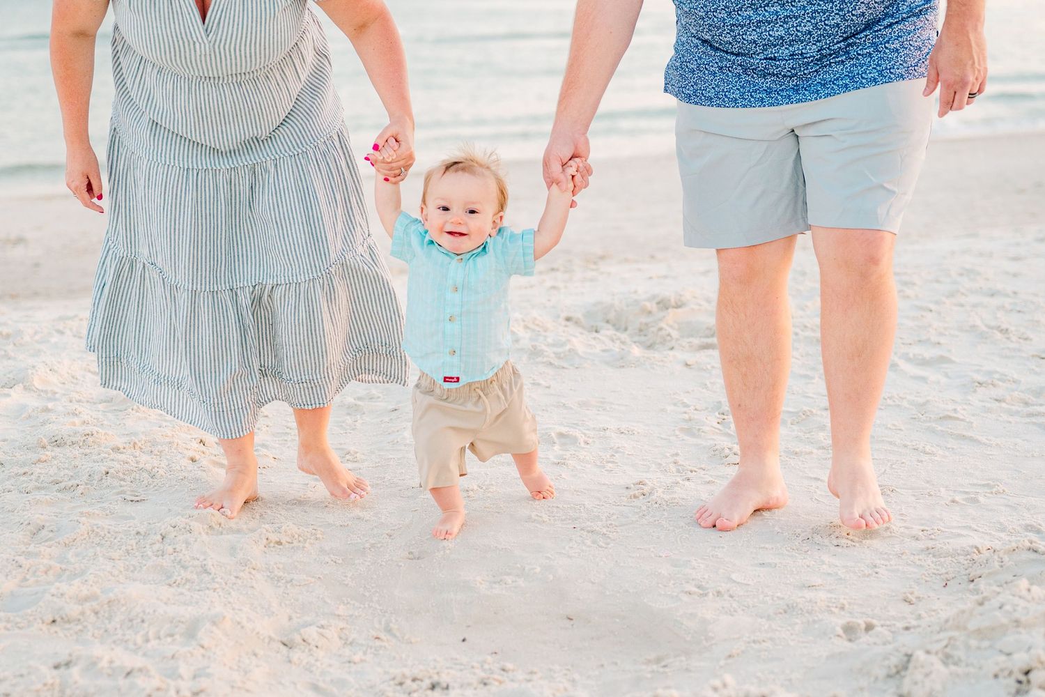 A small child in a blue shirt being lifted by their hands while walking on a sandy beach with two adults.