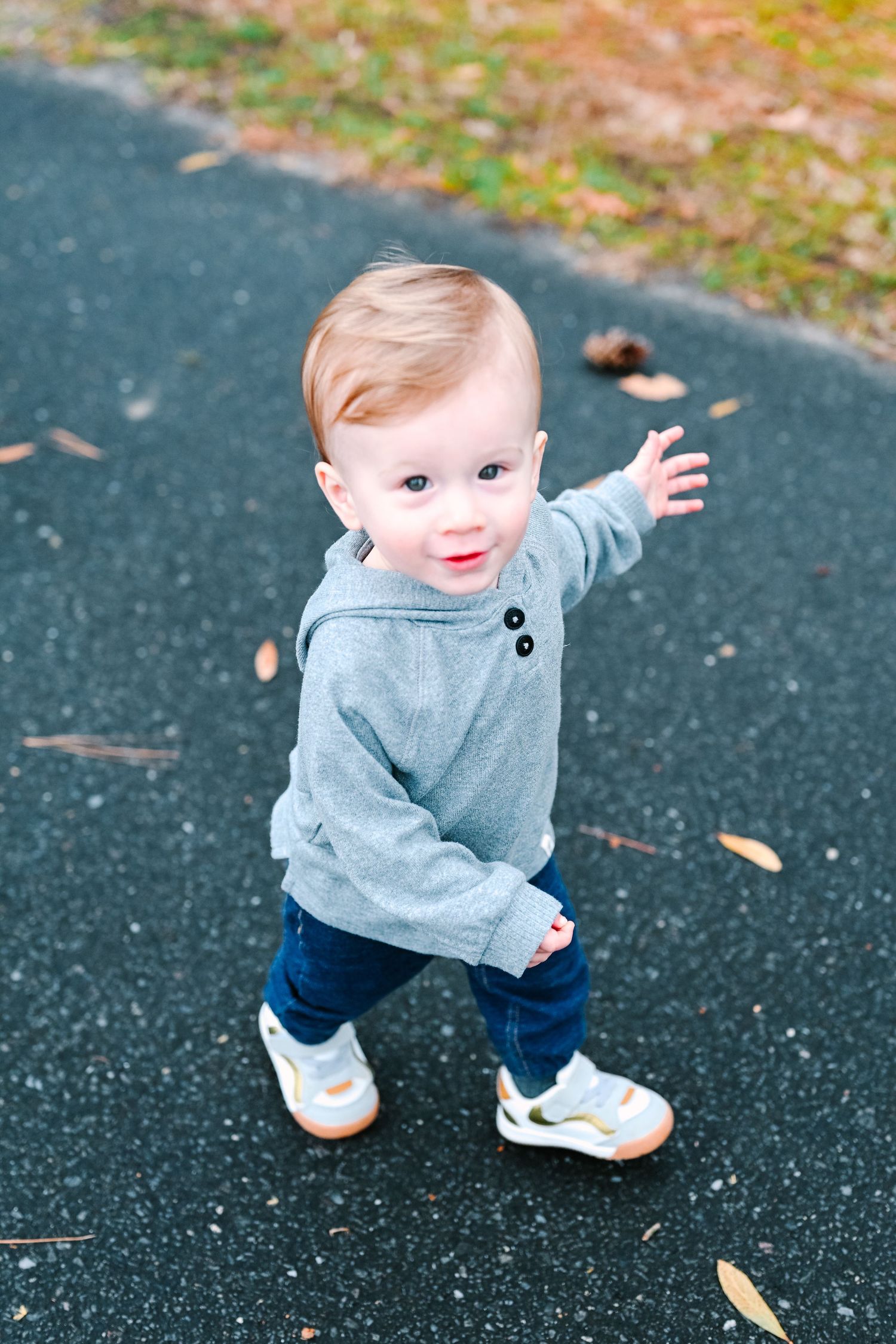 Young toddler in grey sweatshirt and jeans reaches out while walking on dark pavement with autumn leaves scattered around.