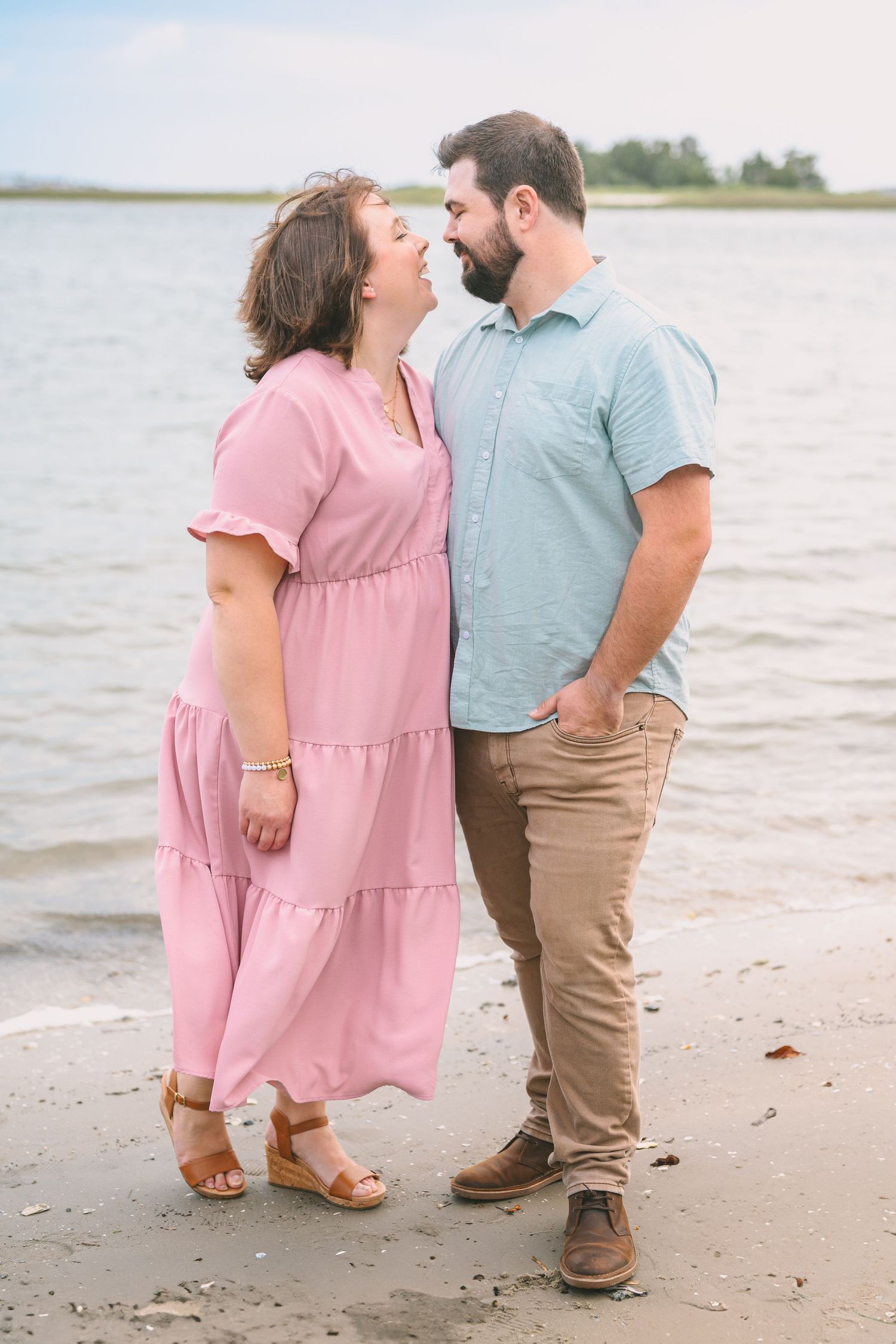 A romantic beachside moment between a couple wearing pink and blue, standing on the sand near calm waters.