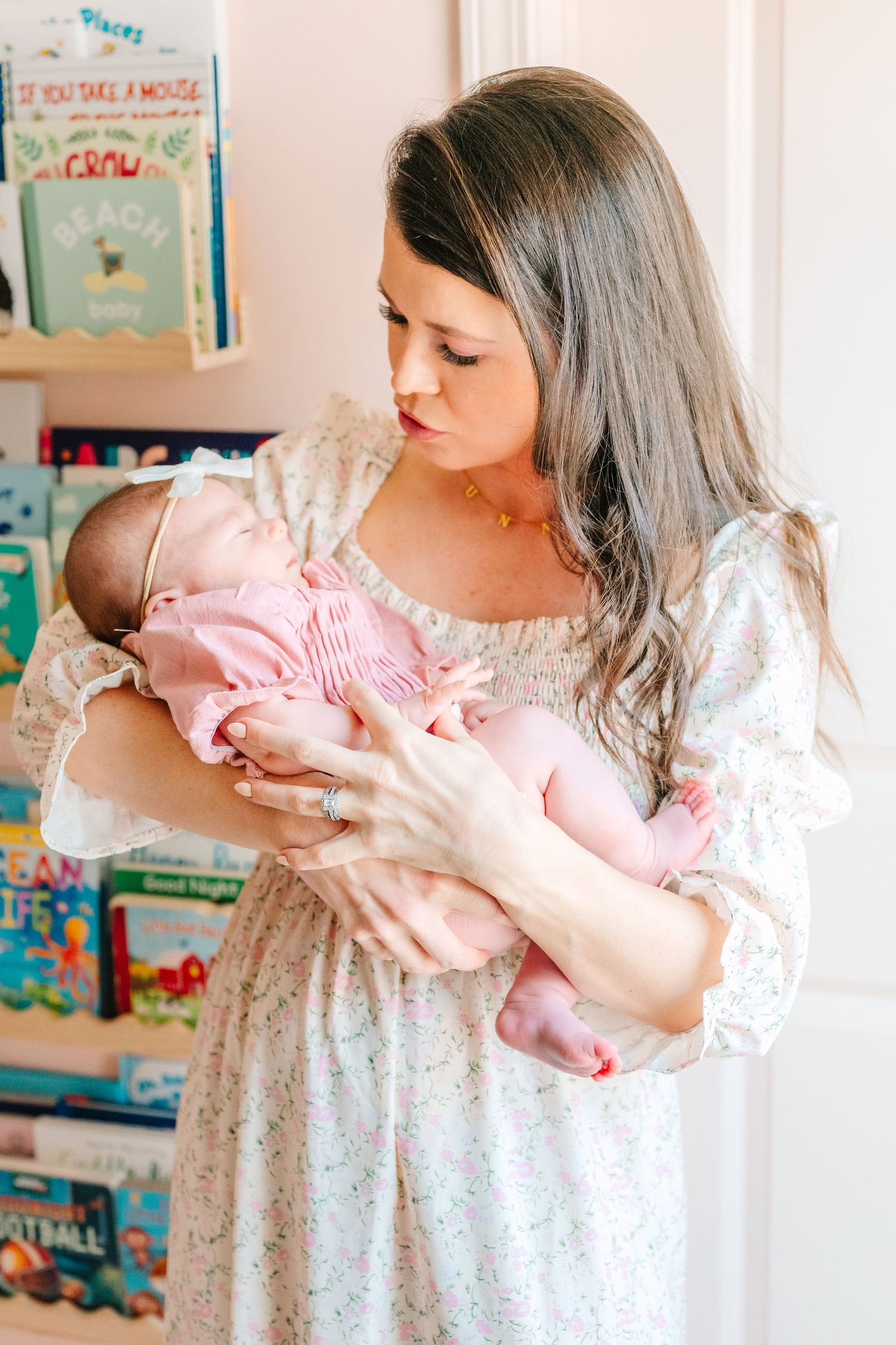 A tender moment captured as a mother in a floral dress holds and gazes down at an infant wearing pink in a nursery setting.