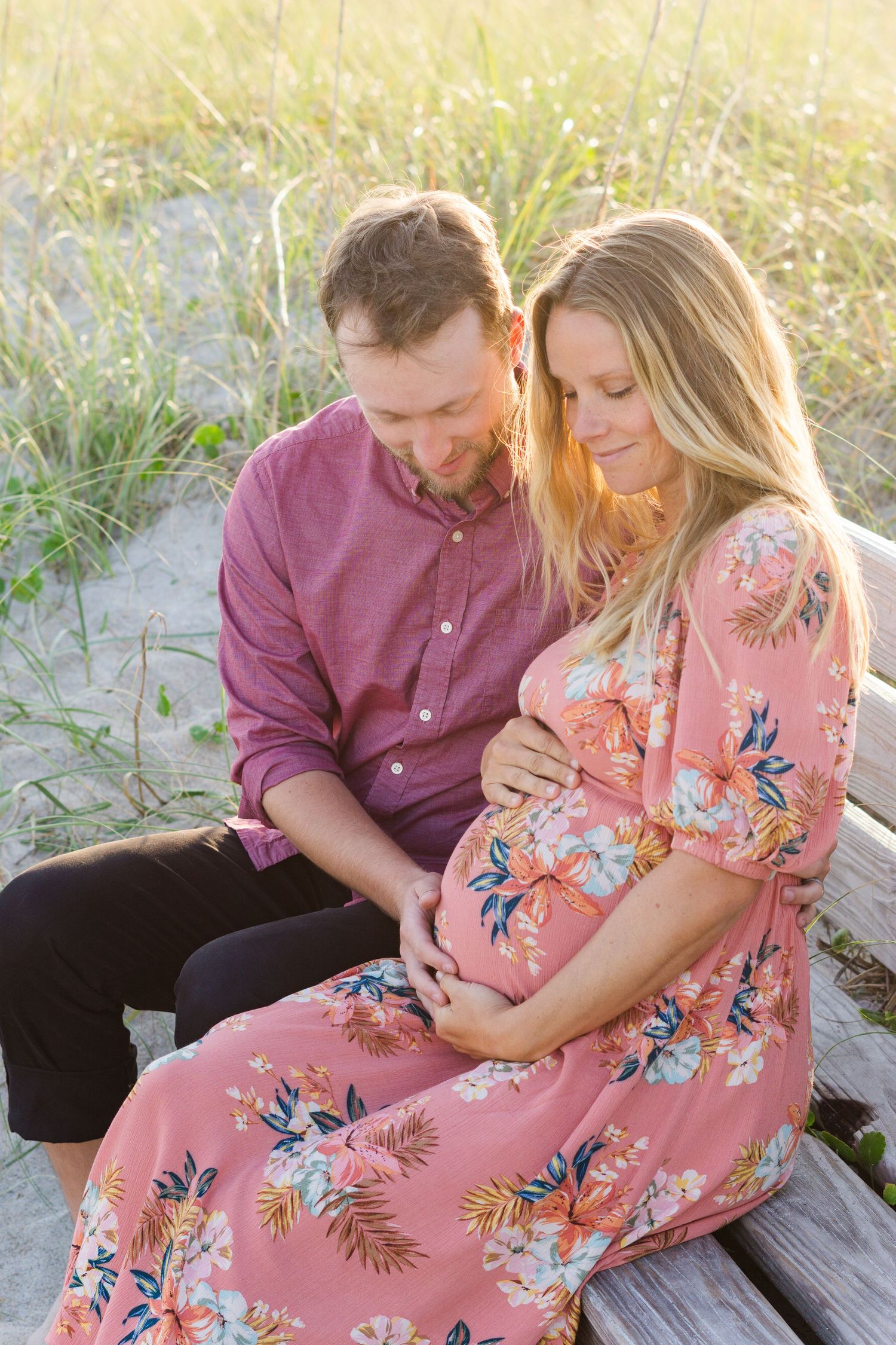 Couple sits on beach during sunset as they lovingly embrace a baby bump in matching pink outfits.