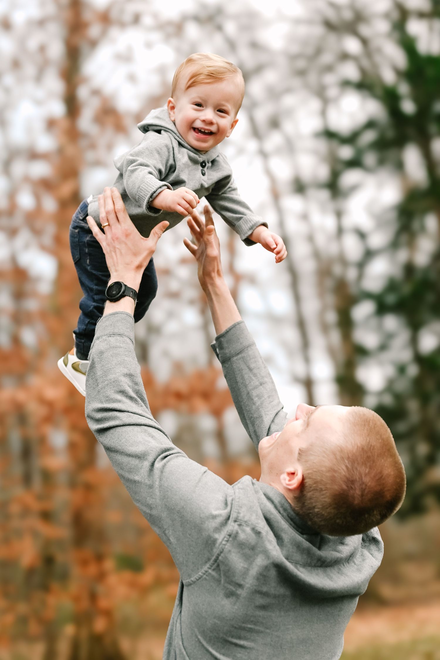 A joyful moment captured outdoors as child is lifted high in the air, both wearing grey sweaters against an autumn backdrop.