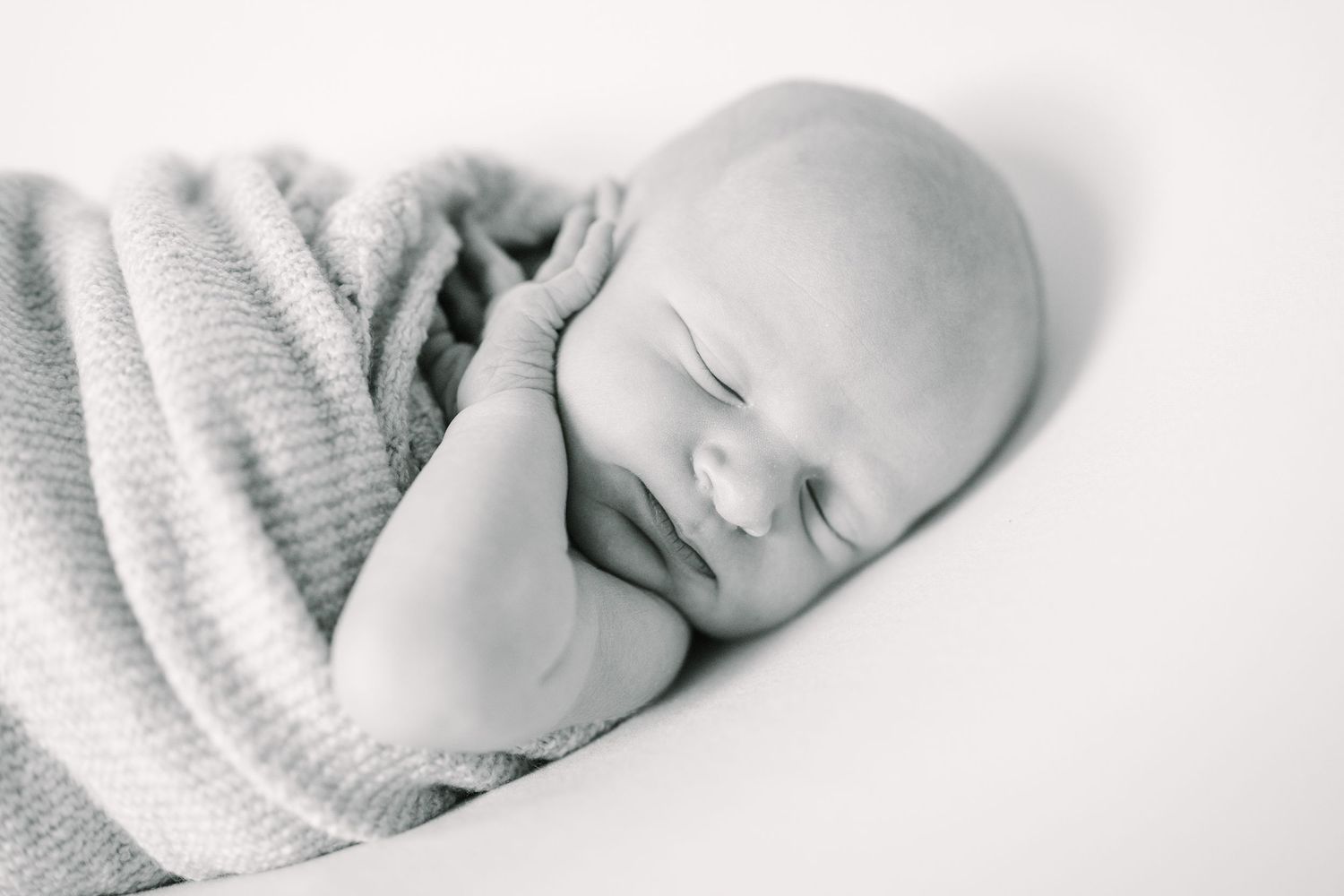 Black and white image of sleeping newborn swaddled in soft gray blanket against white background.