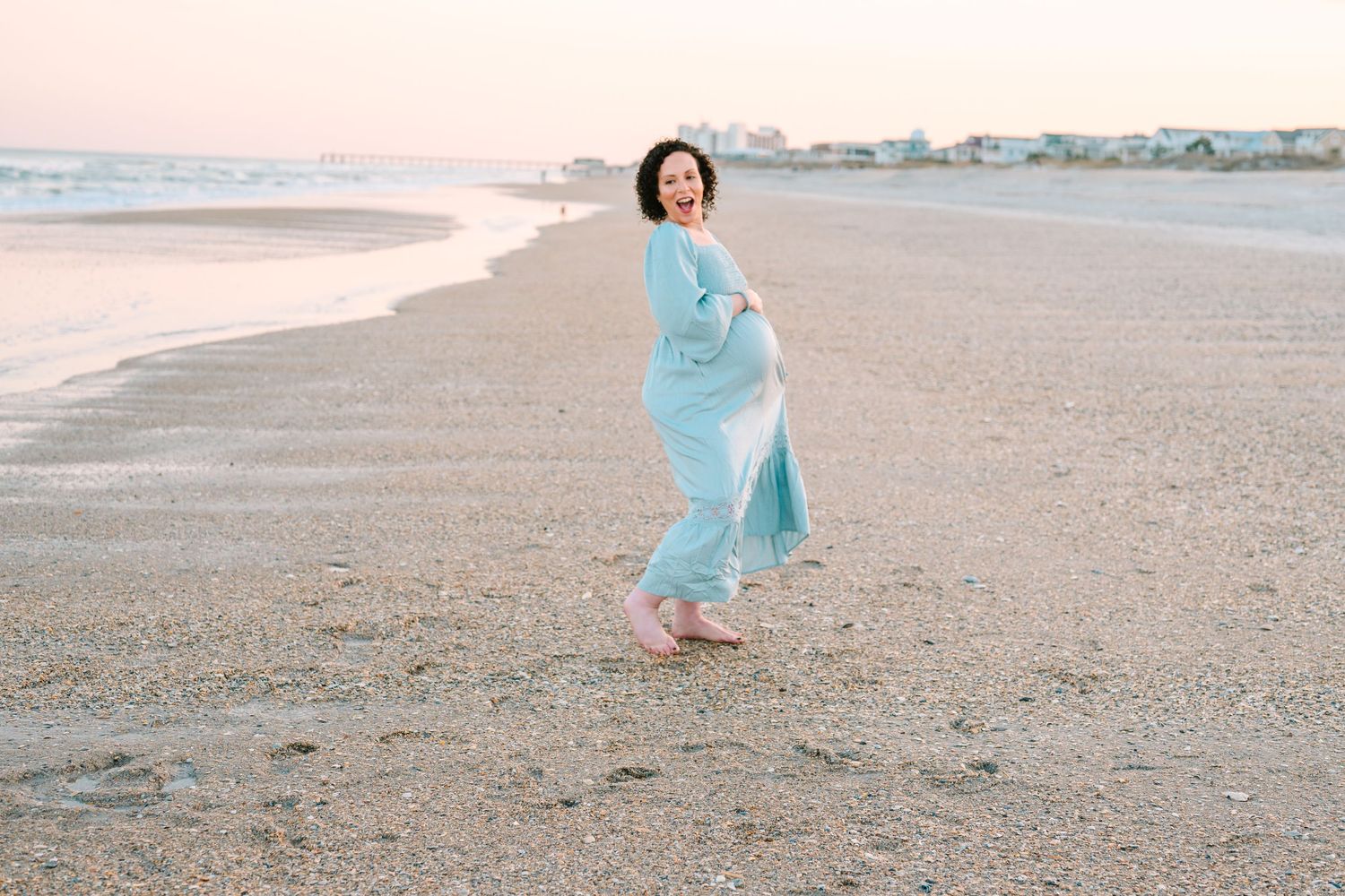 A person in a flowing light blue dress walks gracefully along a peaceful beach at sunset.