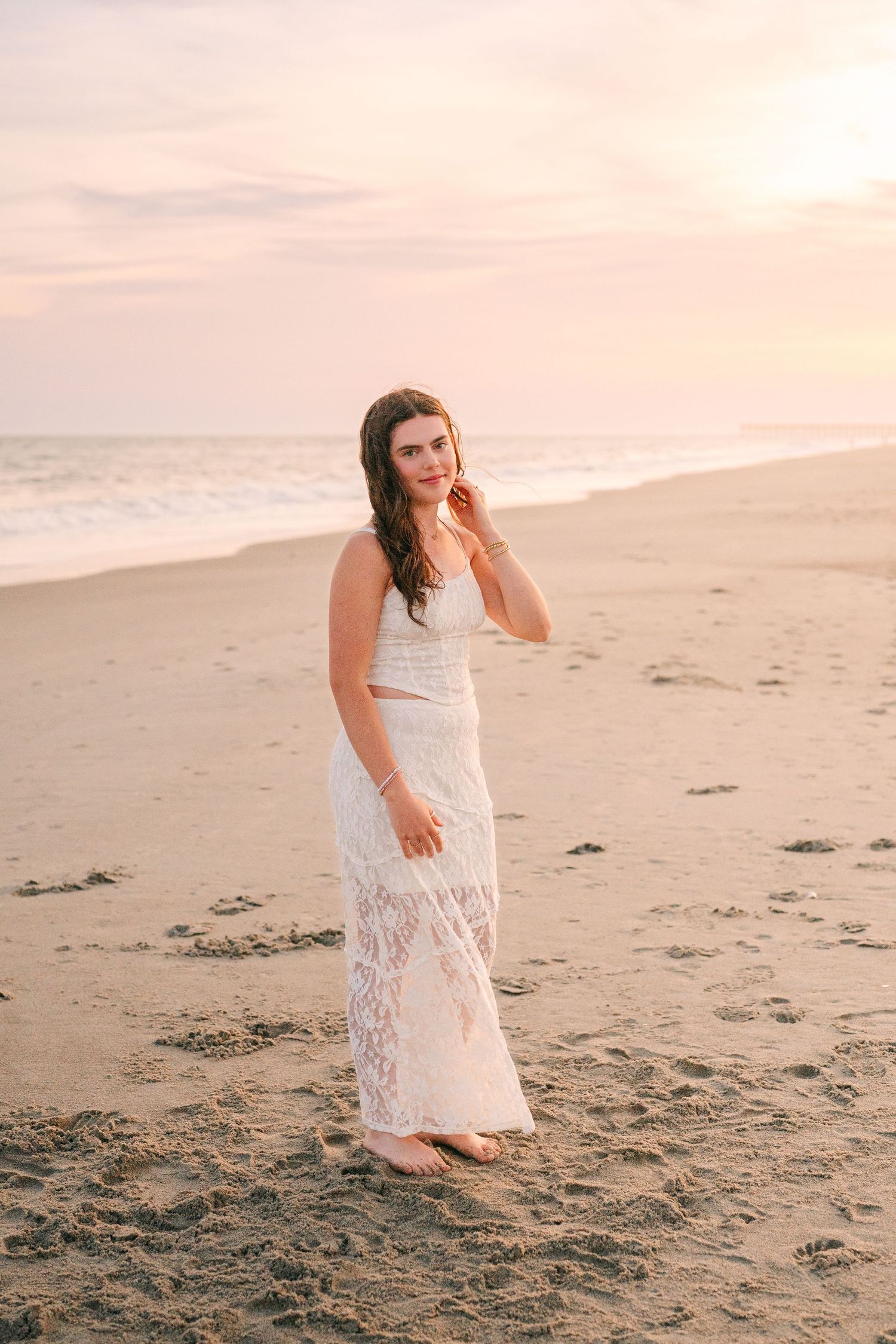 Woman in white lace dress standing alone on beach during golden hour.