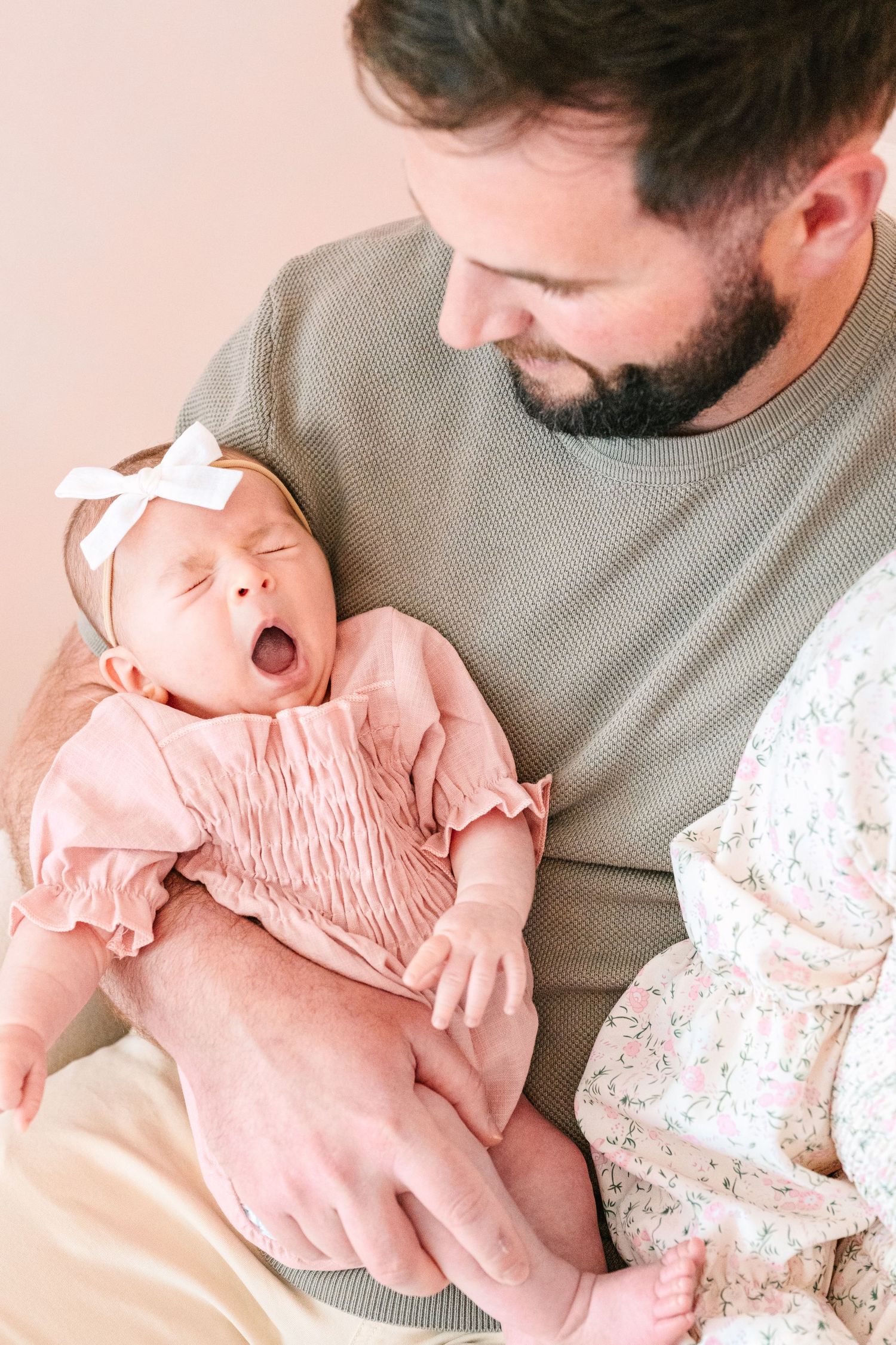 Newborn baby wearing pink smocked outfit yawning while cradled by adult in gray sweater.