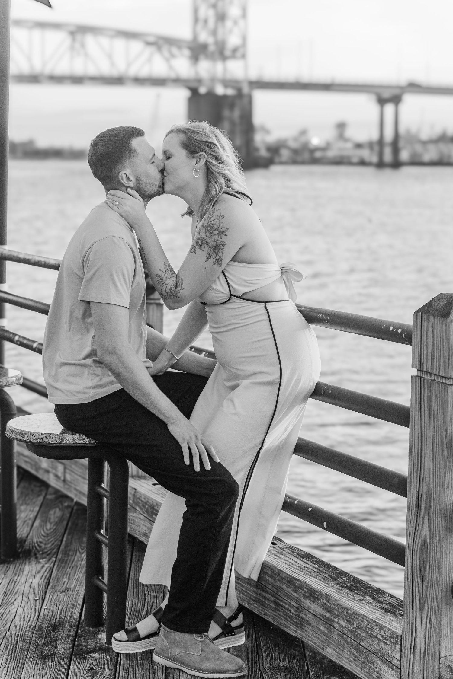 A couple shares a romantic kiss on a wooden pier with a bridge visible in the background.