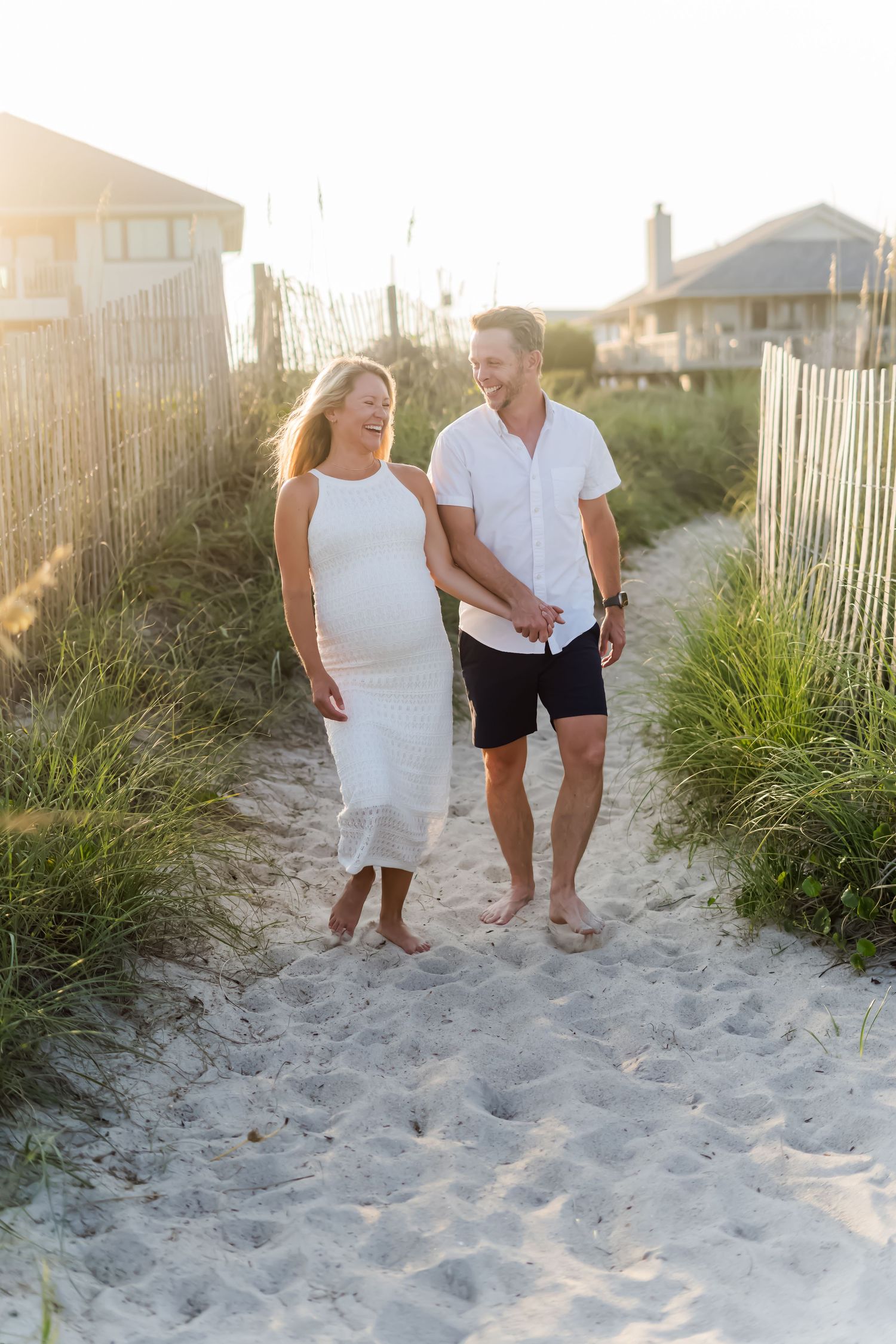 Couple strolls together on sandy coastal pathway between beach fences at sunset.