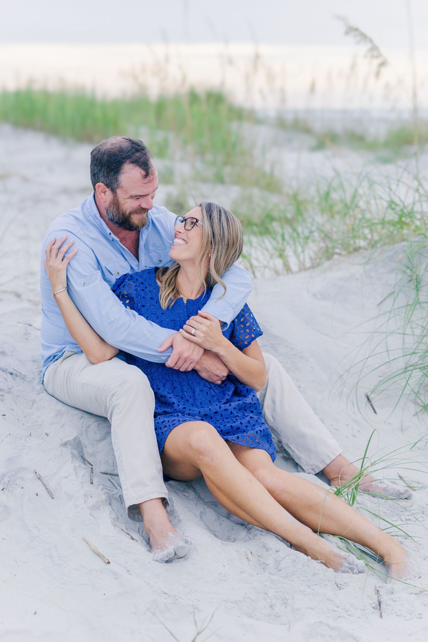Couple embraces while sitting on coastal sand dunes with beach grass in the background.