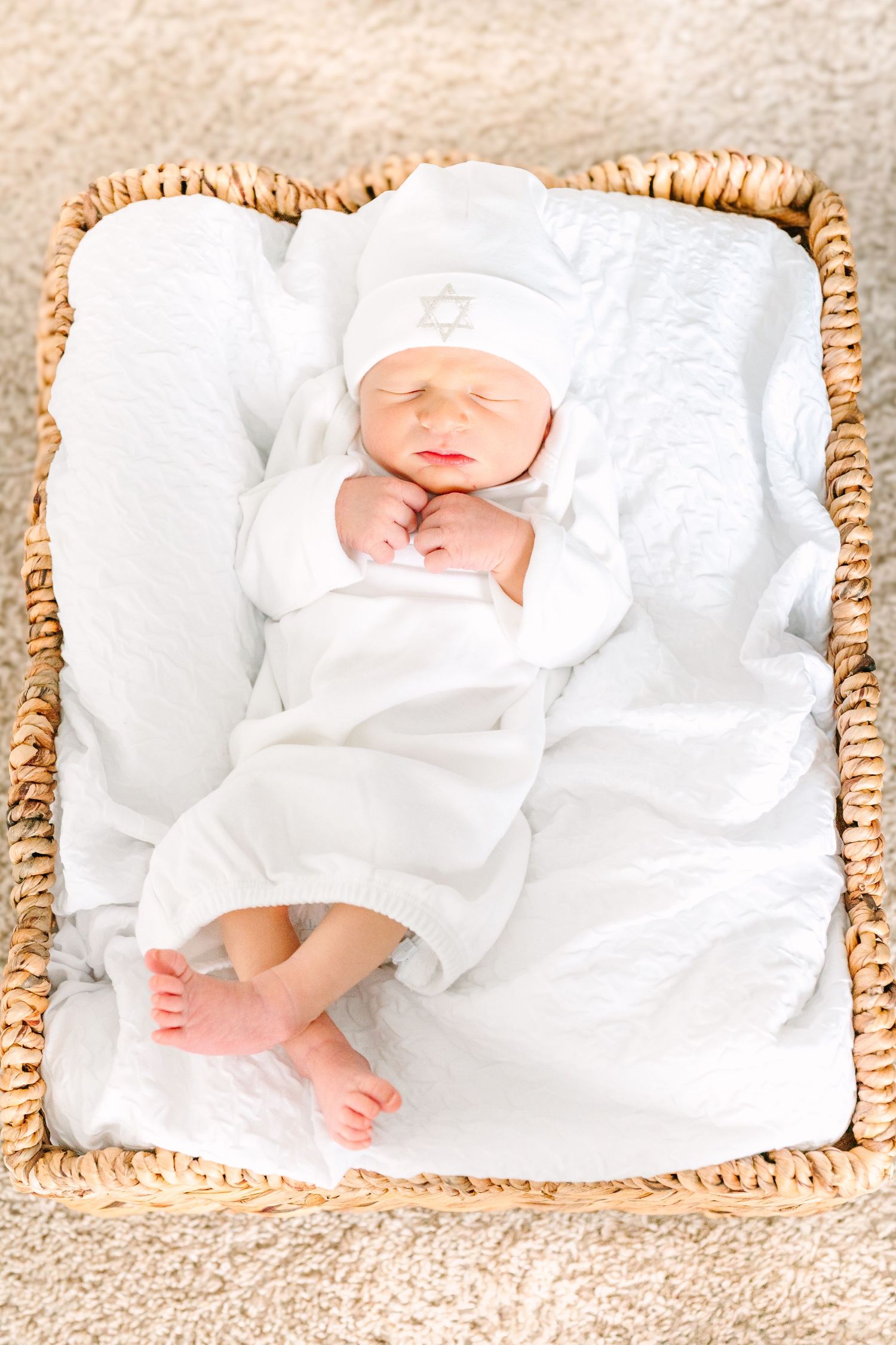Sleeping newborn in white swaddle and hat rests peacefully in woven basket with white blanket.