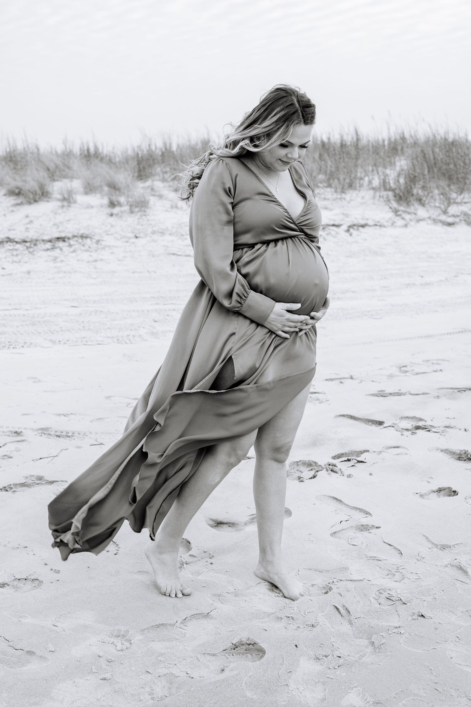 Black and white maternity photo of an expectant mother in a flowy dress on a windswept beach.