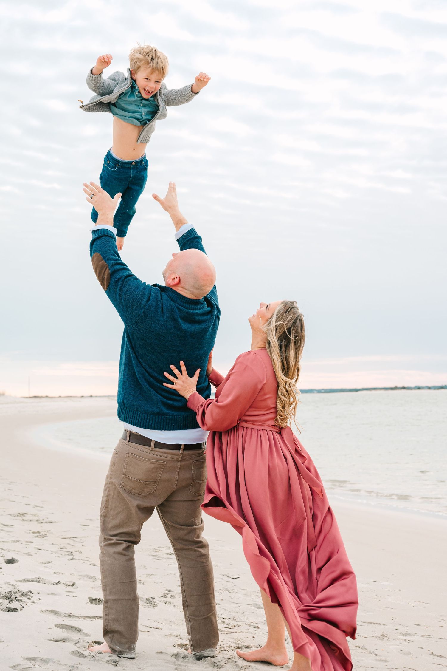 A family plays together at the beach as the parent lifts a small child high into the air at sunset.
