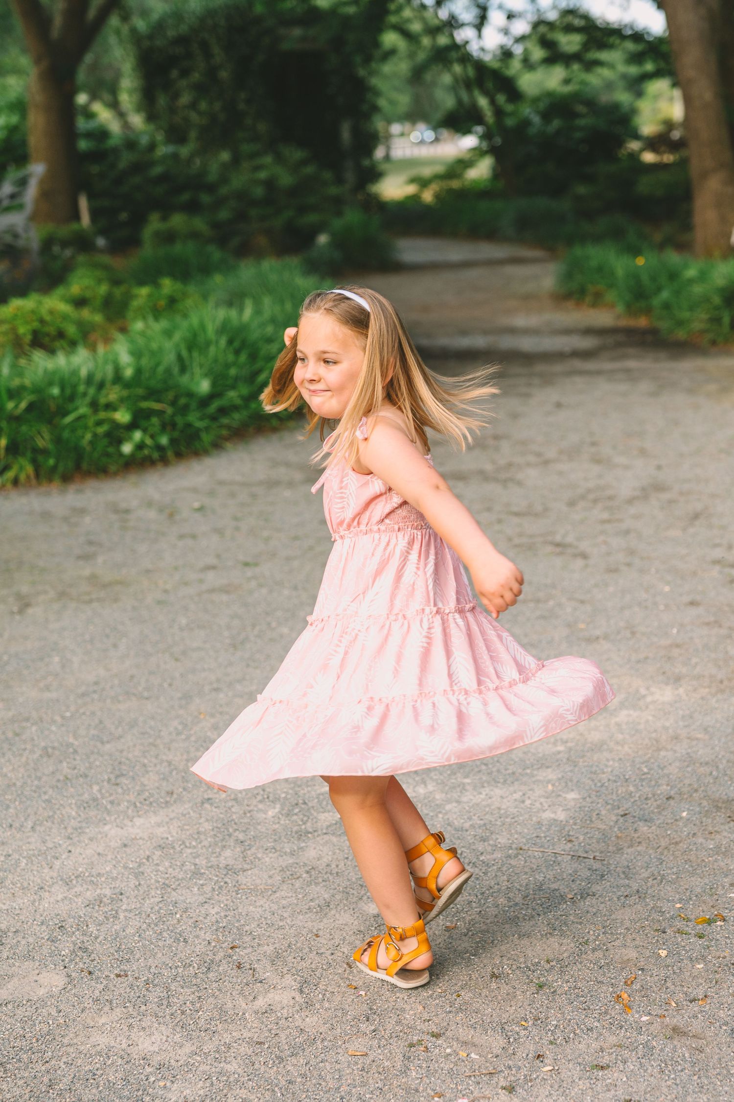 Someone in a pink tiered dress twirls joyfully on a tree-lined garden path while wearing brown sandals.