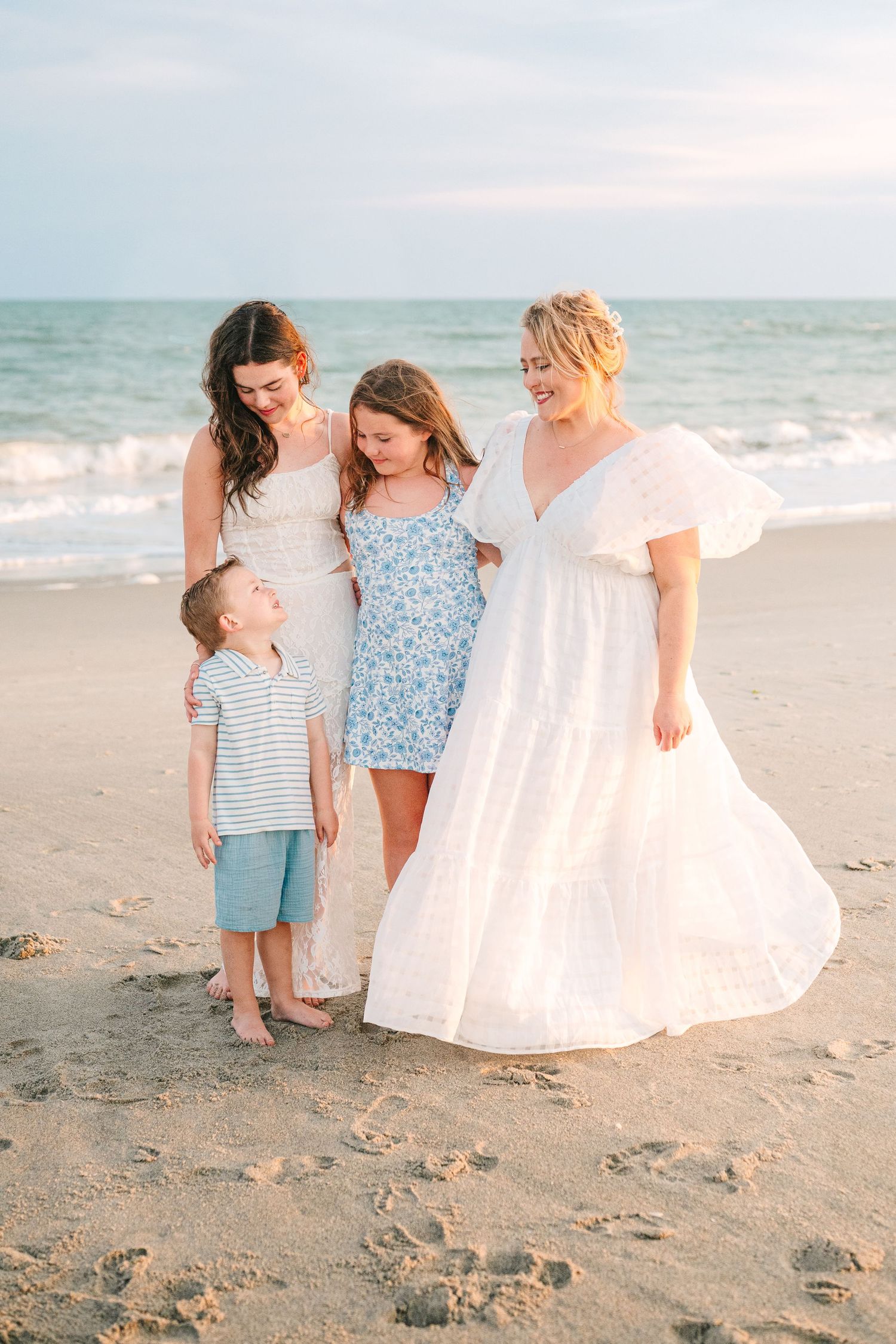 Small group in white and blue dresses standing together on beach at sunset.