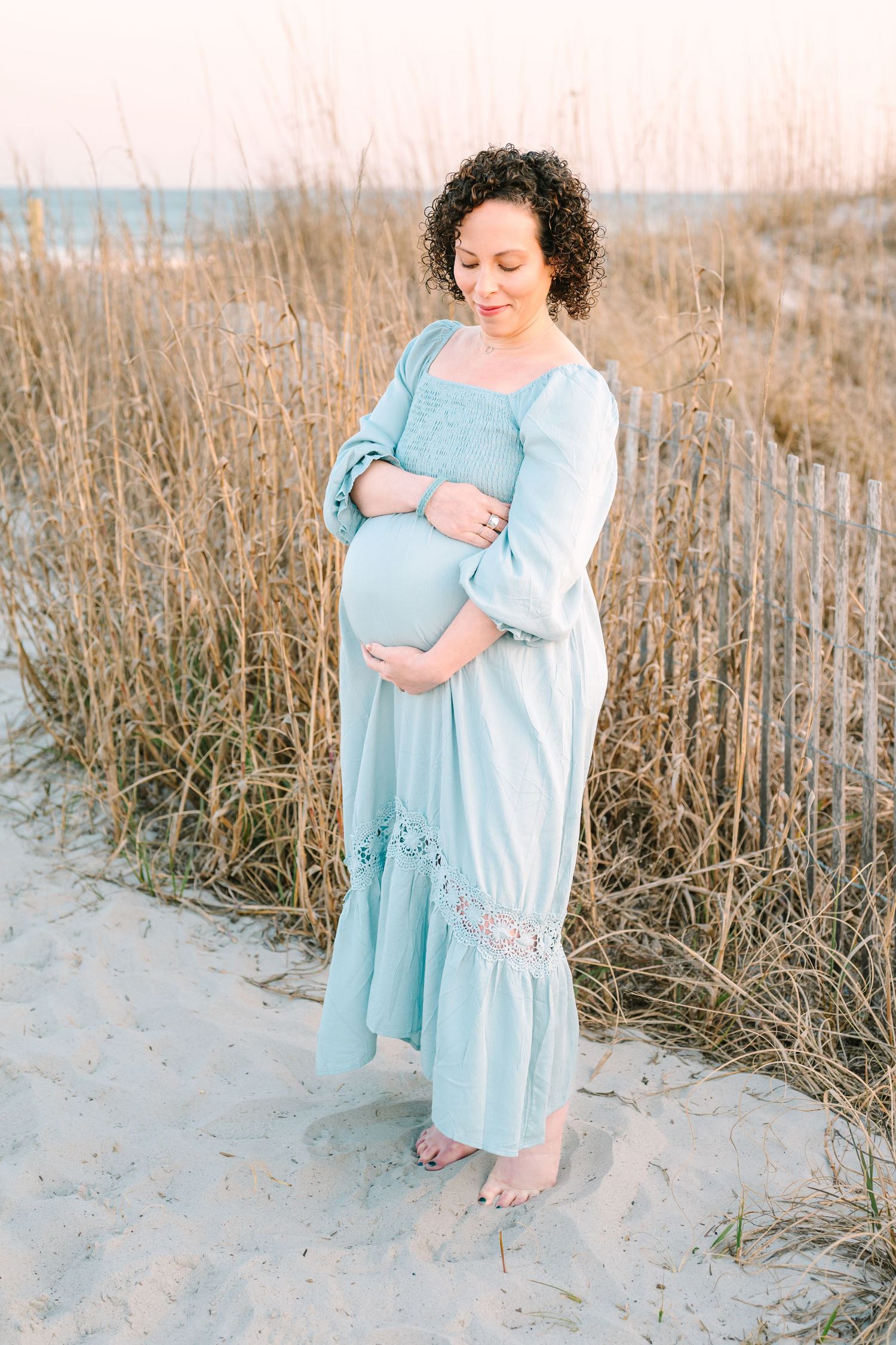 A pregnant person in a flowing light blue dress stands barefoot on sandy beach dunes with tall grass in the background.
