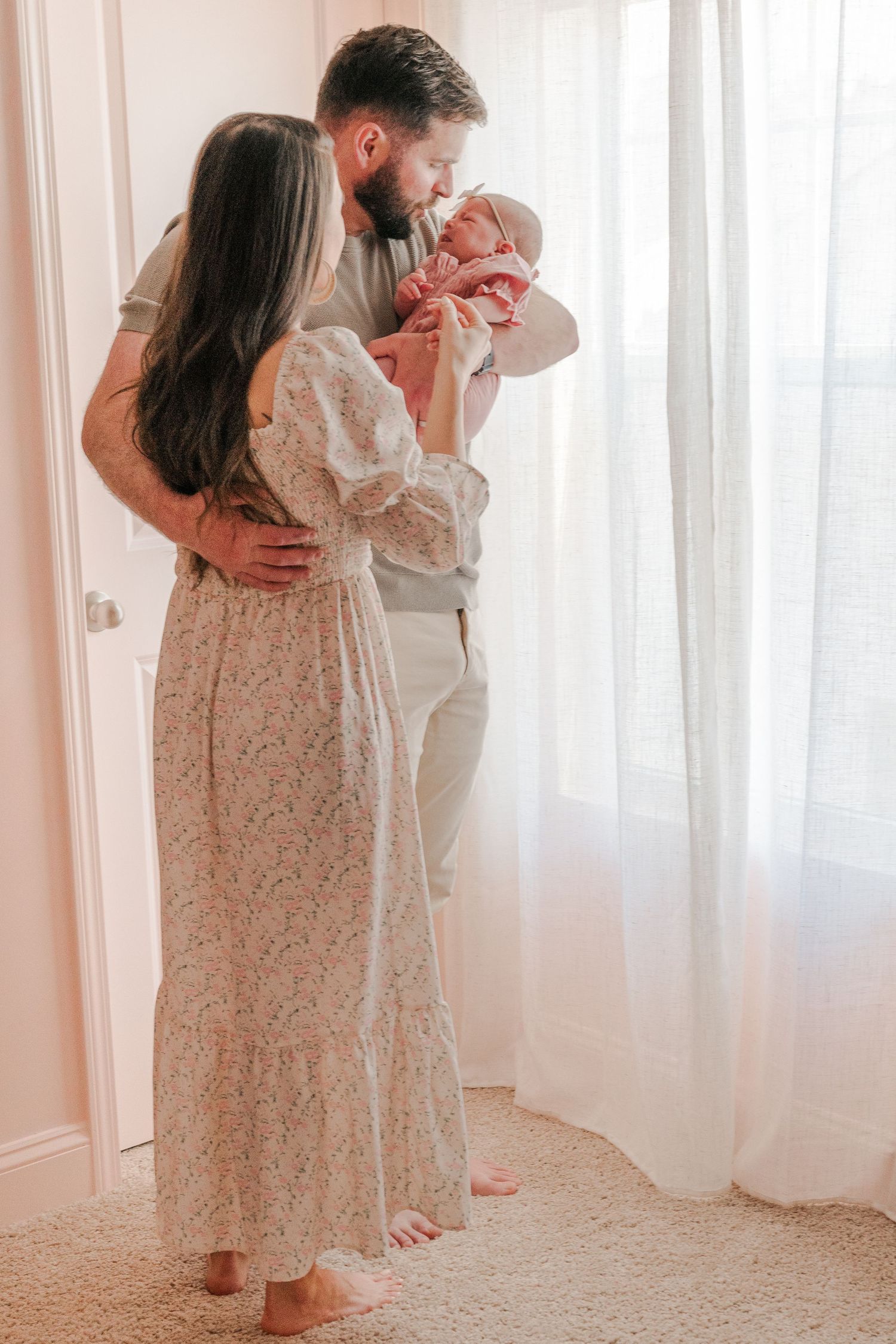 A couple in a sunlit nursery sharing an intimate moment with their newborn baby near sheer white curtains.