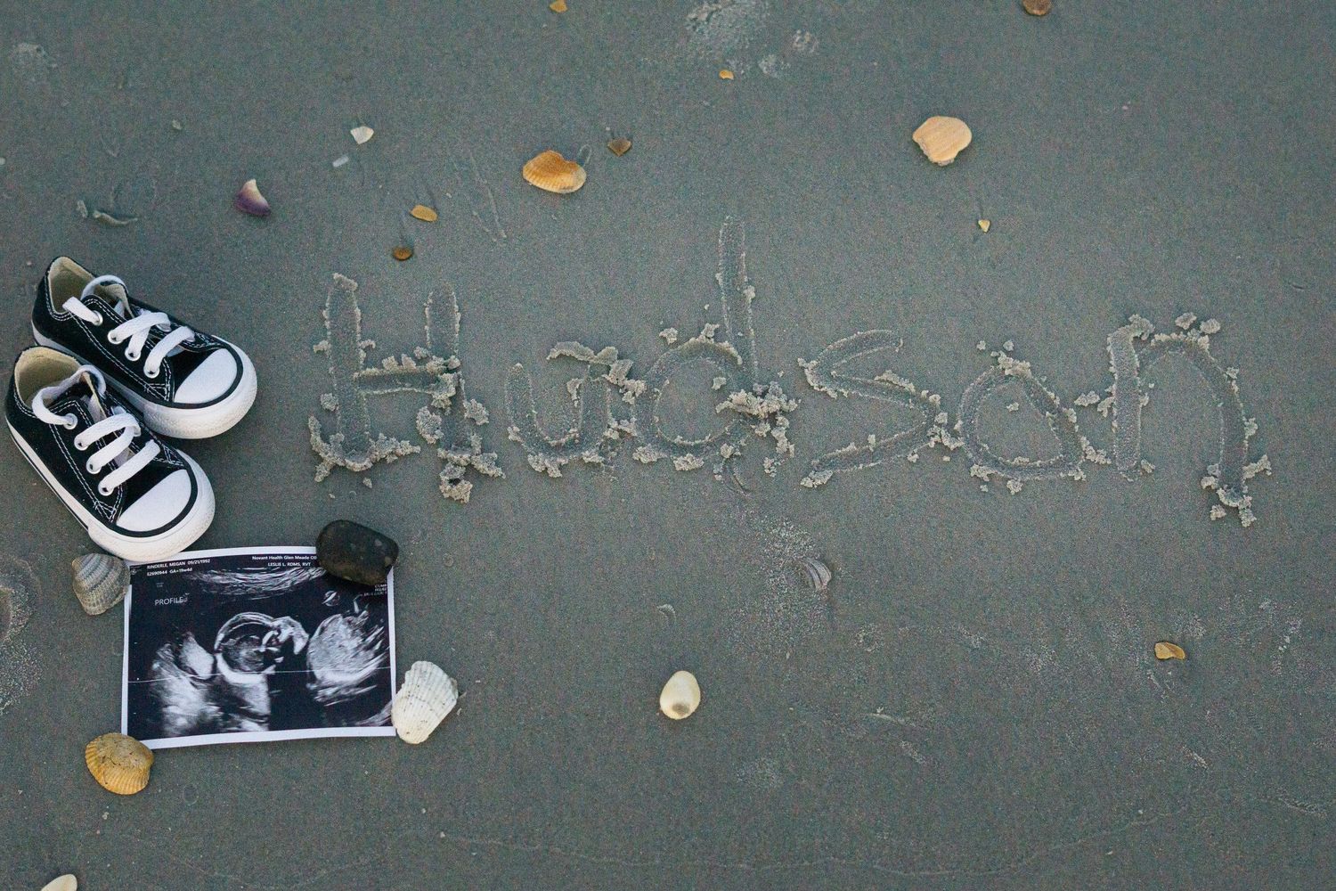 Baby shoes and ultrasound photo next to pregnancy announcement written in beach sand.