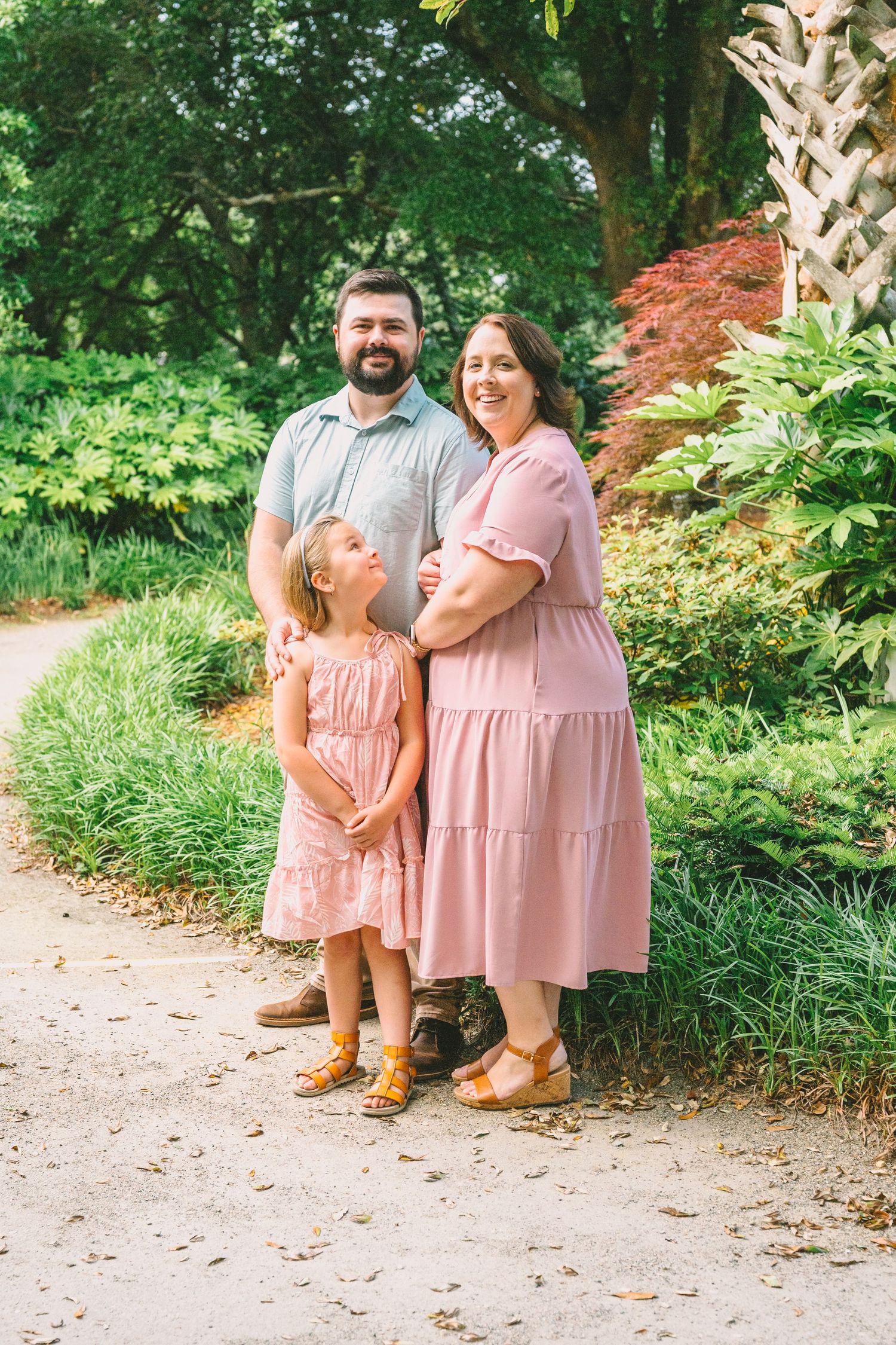 A family wearing pink and blue casual outfits poses together on a garden pathway surrounded by greenery.