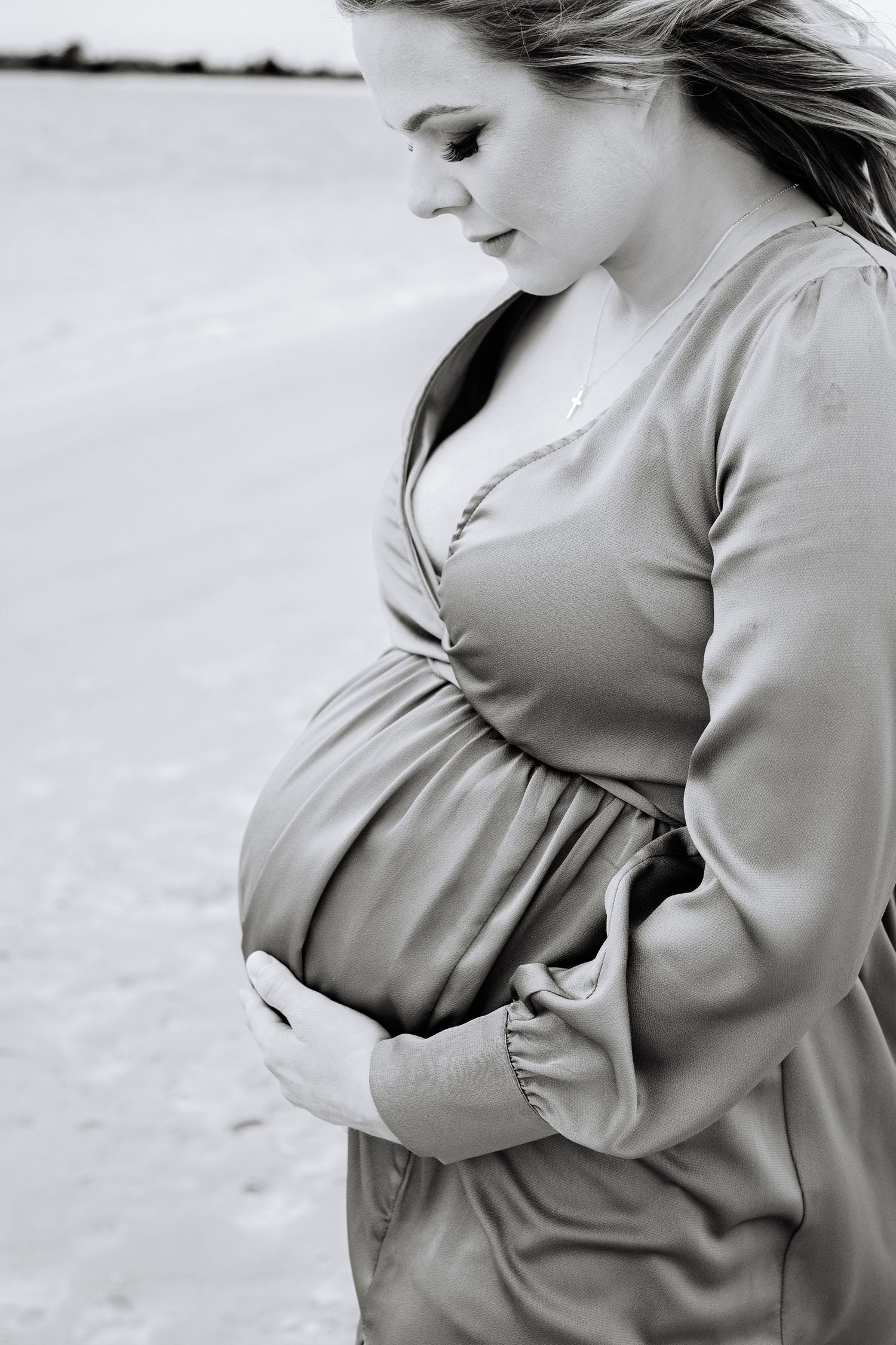 Black and white portrait of an expectant mother in a flowing dress tenderly holding her pregnant belly outdoors.