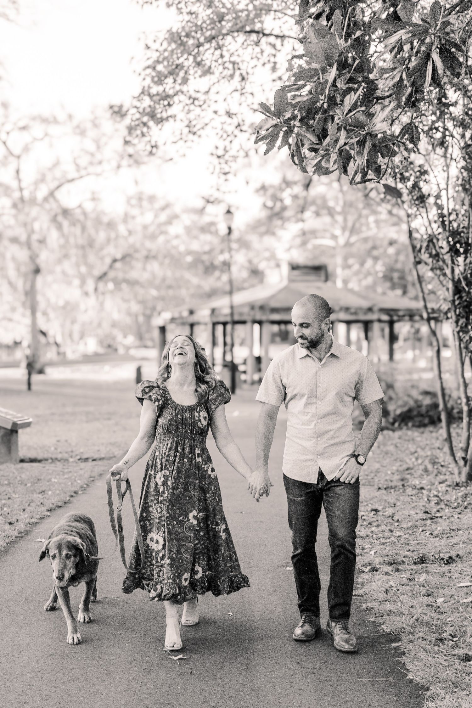 A couple walks along a park path holding hands with their dog on a sunny day.
