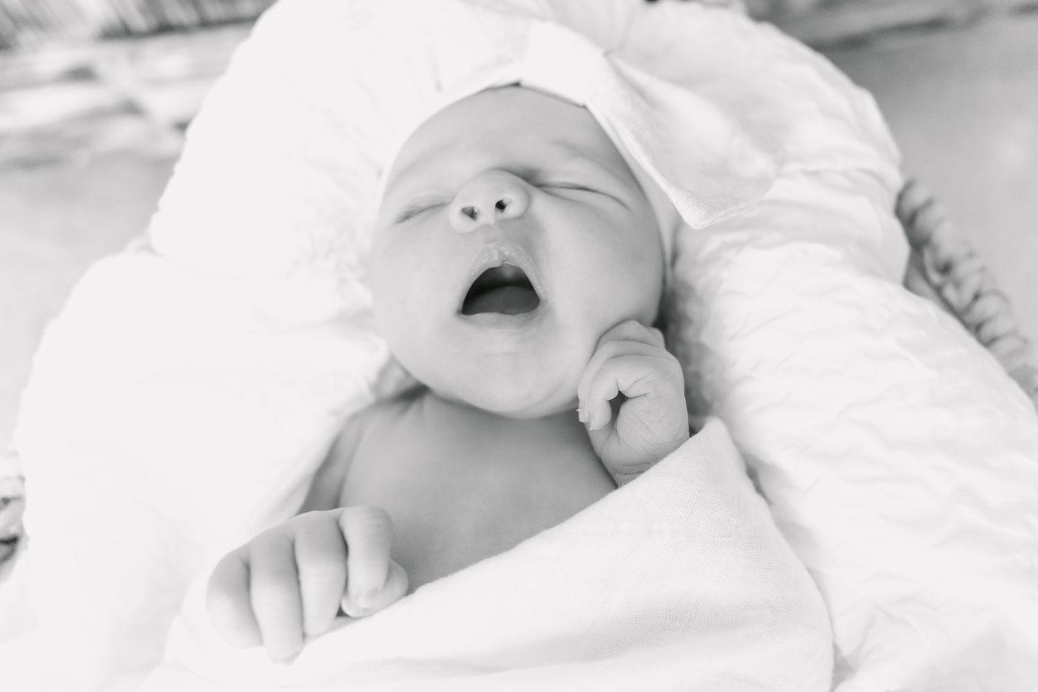 Black and white portrait of yawning newborn baby wrapped in white blanket.